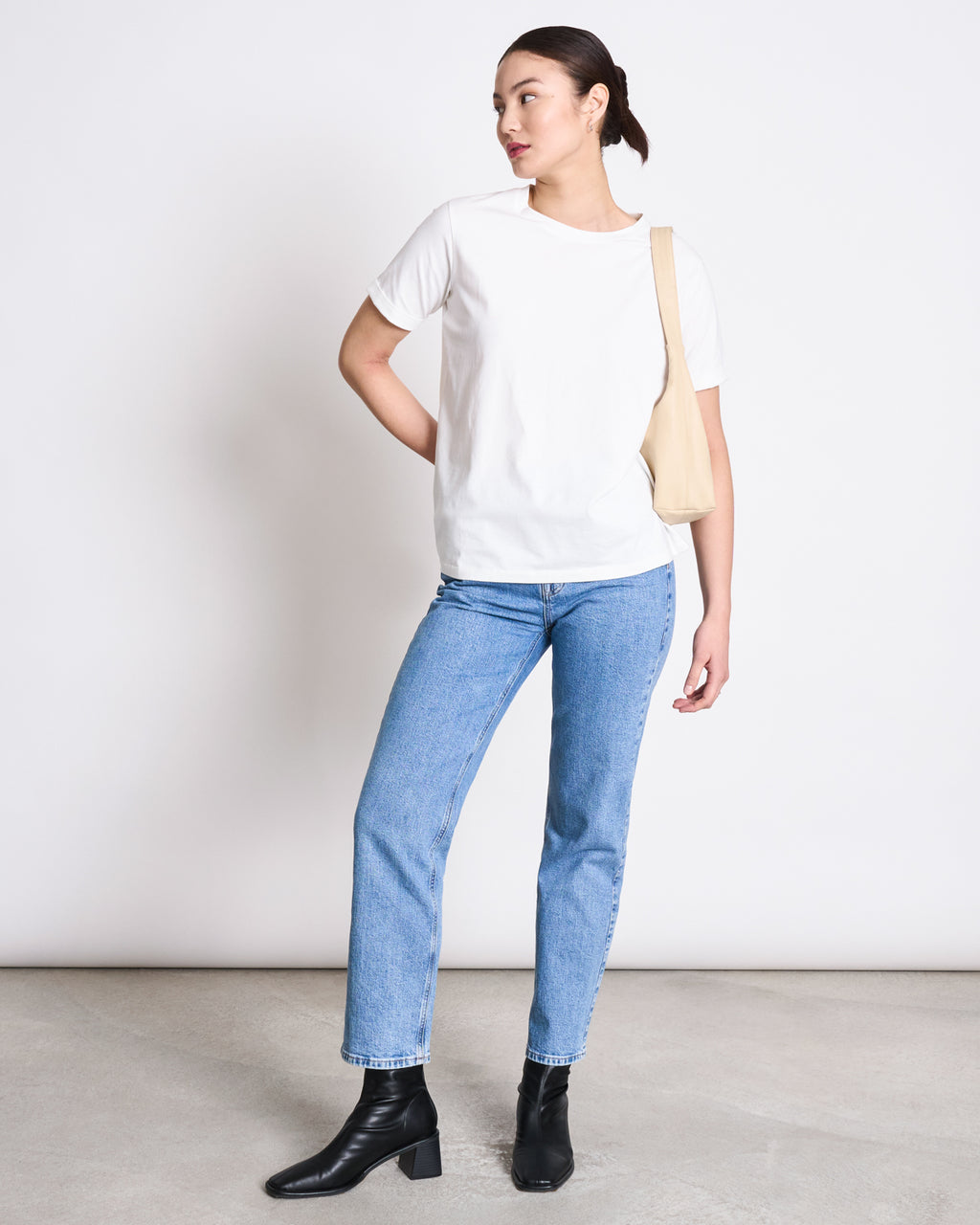 A person stands against a plain white wall, wearing the jan-n-june BRUSHED T-SHIRT BOY PLAIN WHITE GOTS, light blue jeans, black ankle boots, and a cream-colored shoulder bag. Their hair is tied back as they look to the side.