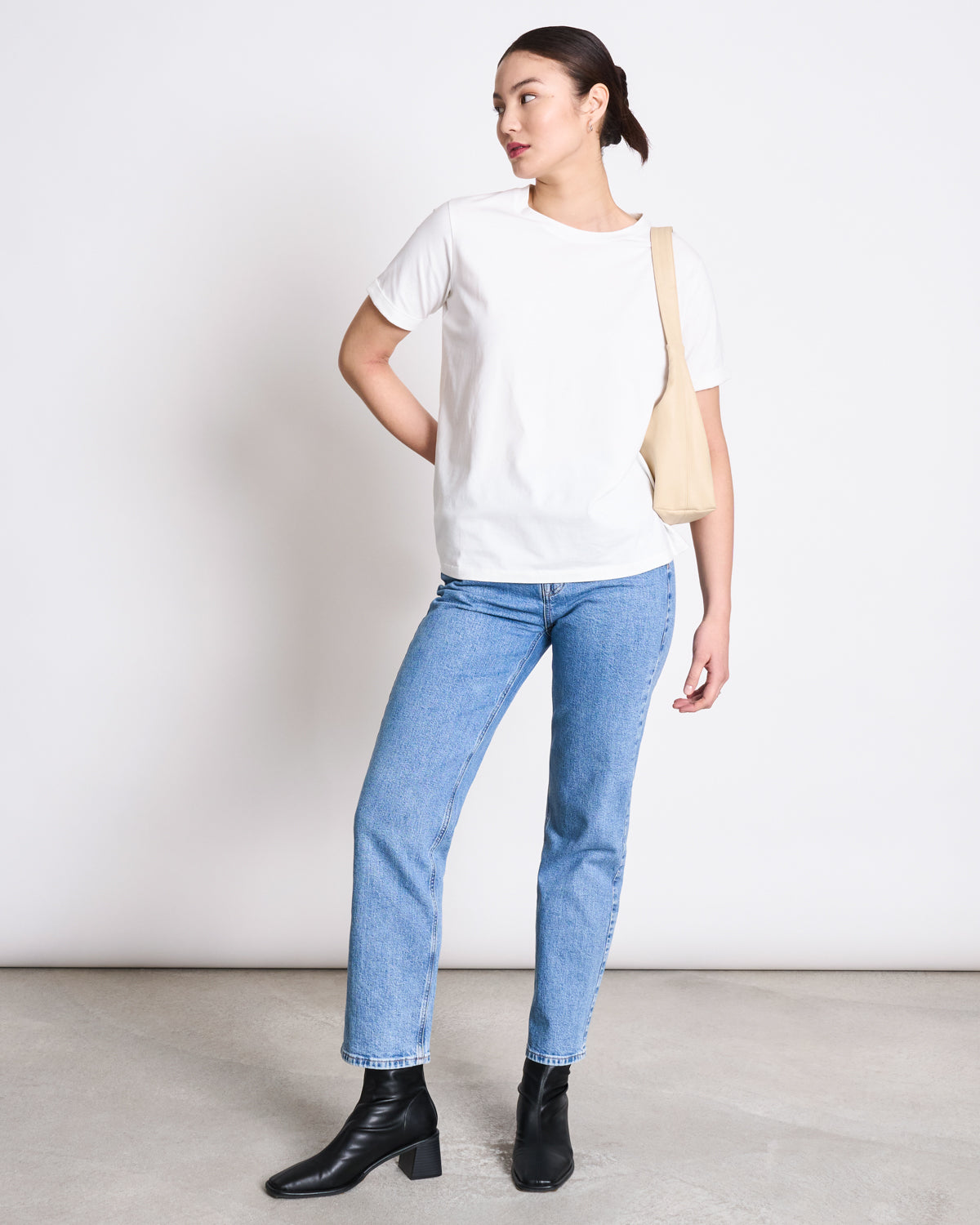 A person stands against a plain white wall, wearing the jan-n-june BRUSHED T-SHIRT BOY PLAIN WHITE GOTS, light blue jeans, black ankle boots, and a cream-colored shoulder bag. Their hair is tied back as they look to the side.