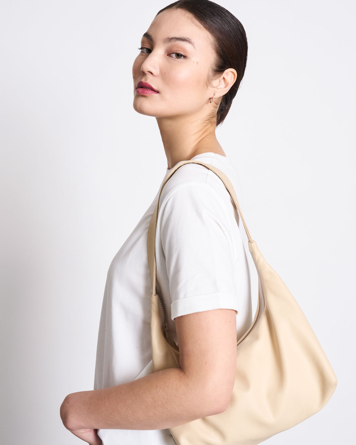A woman with straight dark hair in a low bun wears the BRUSHED T-SHIRT BOY PLAIN WHITE GOTS by jan-n-june and holds a beige shoulder bag, posing against a light background and looking over her shoulder at the camera.
