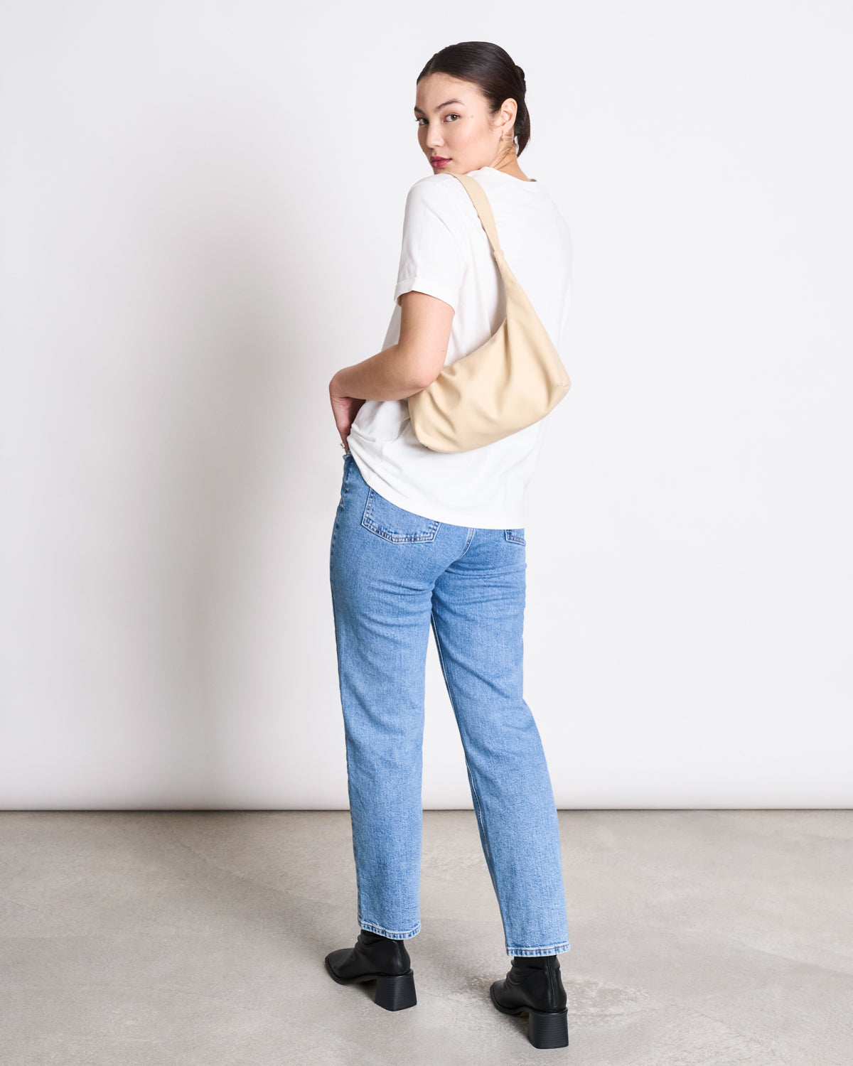 A woman faces away, glancing back over her shoulder. She wears the jan-n-june BRUSHED T-SHIRT BOY PLAIN WHITE GOTS with light blue jeans, black shoes, and a beige shoulder bag against a plain light background.
