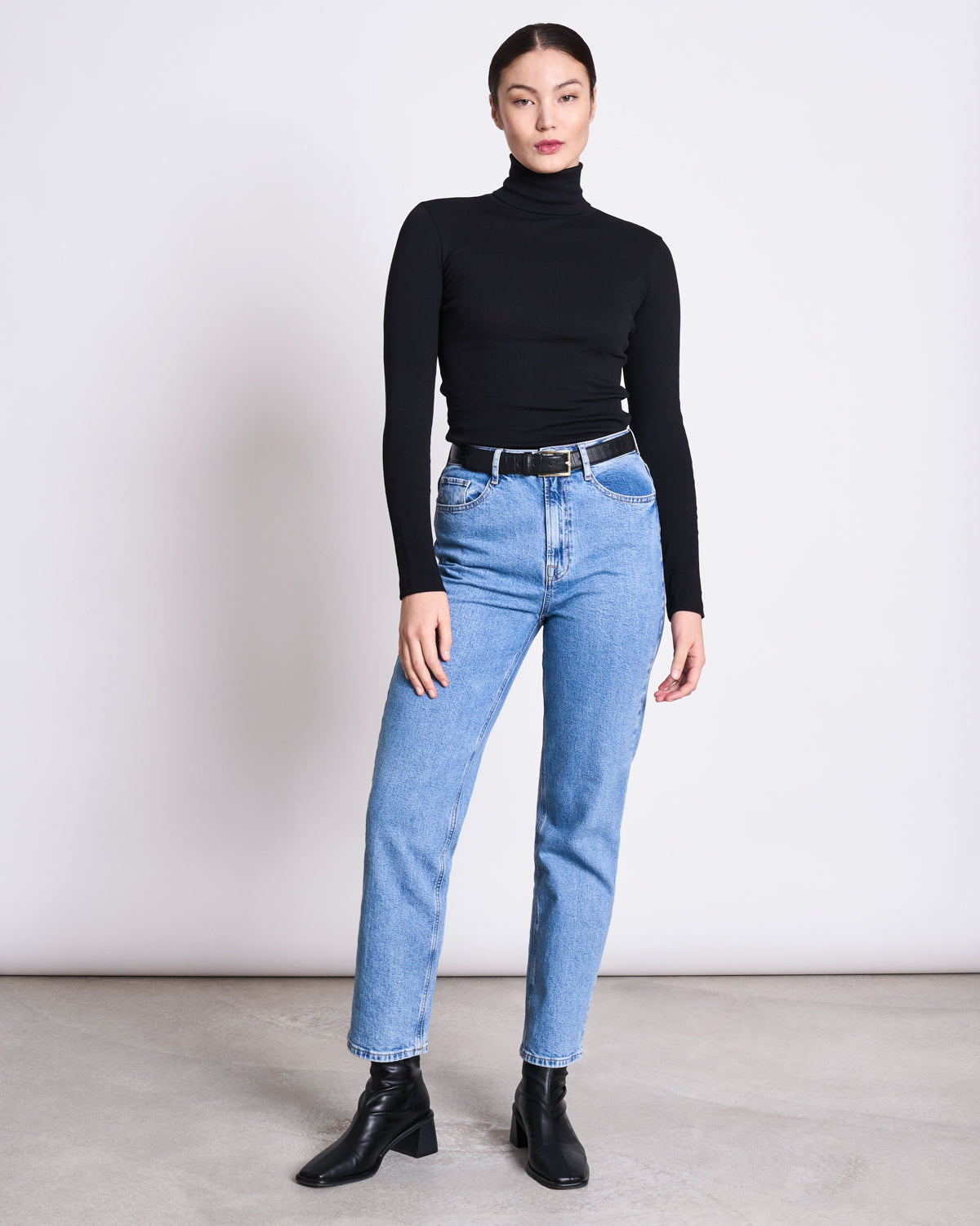 A person stands against a plain background, wearing the jan-n-june MINIRIB TURTLENECK CHUVA BLACK GOTS, light blue high-waisted jeans, a black belt, and black ankle boots. Their straight dark hair is pulled back.