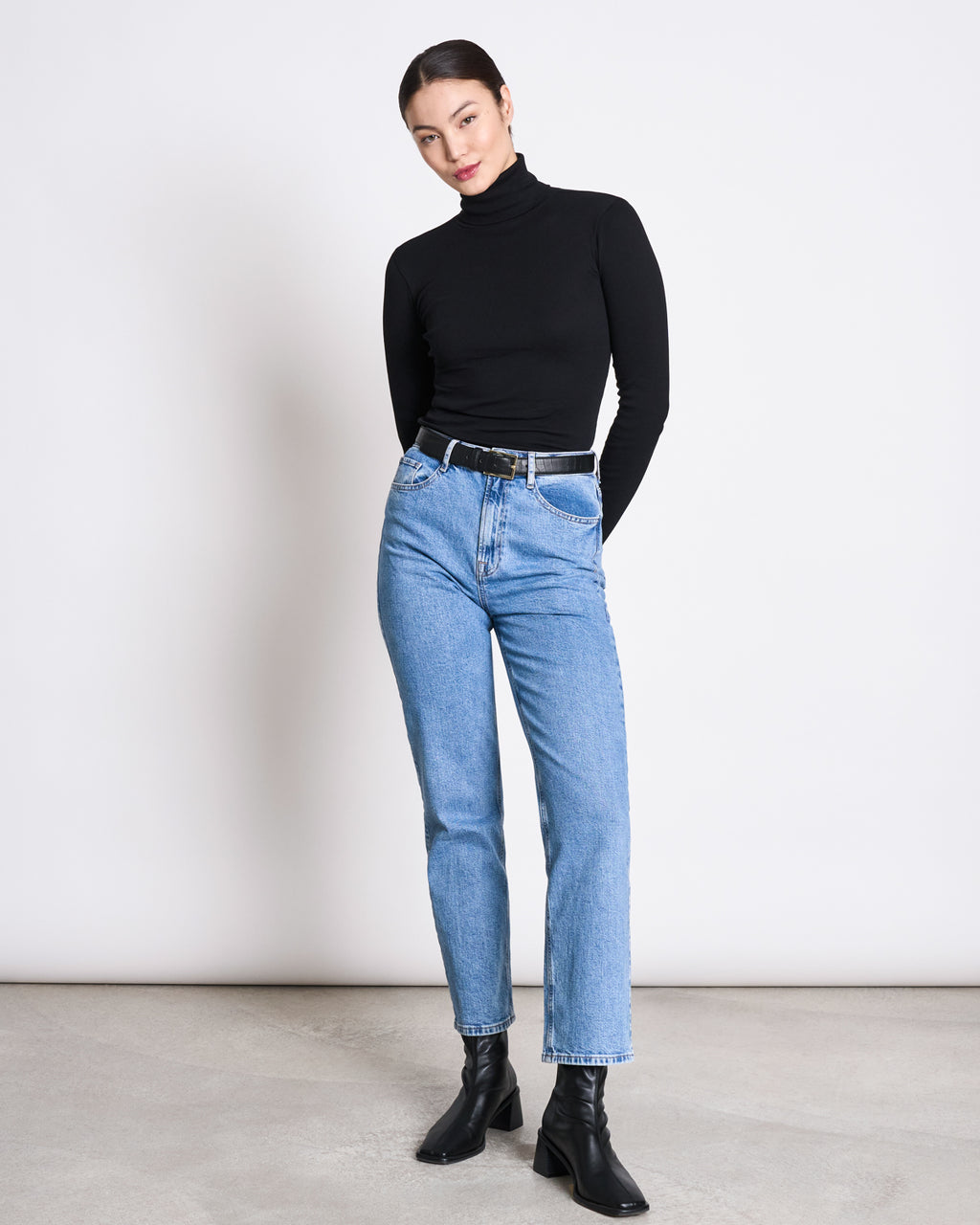 A woman stands against a plain background wearing the jan-n-june MINIRIB TURTLENECK CHUVA BLACK GOTS, light blue high-waisted jeans, a black belt, and black ankle boots. Her hands are behind her back as she looks slightly downward.