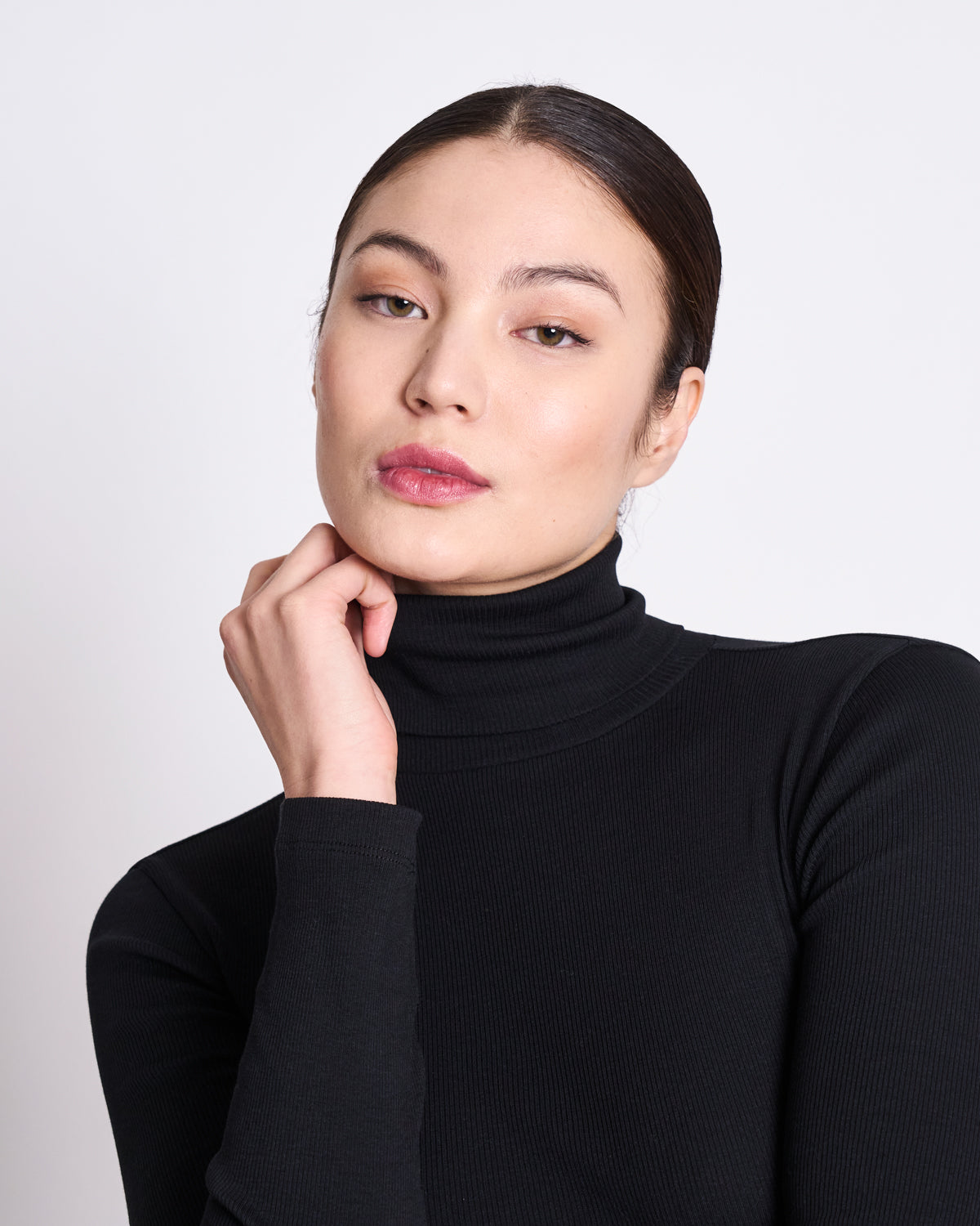 A person with fair skin and dark hair pulled back wears the jan-n-june MINIRIB TURTLENECK CHUVA BLACK GOTS, resting their chin on their hand and looking calmly at the camera against a plain light background.