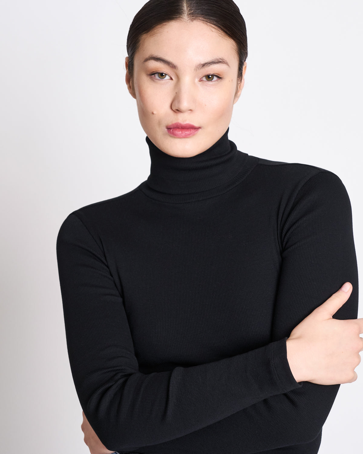 A person with straight dark hair pulled back wears a jan-n-june MINIRIB TURTLENECK CHUVA BLACK GOTS, standing against a plain white background with arms loosely crossed and a neutral expression.