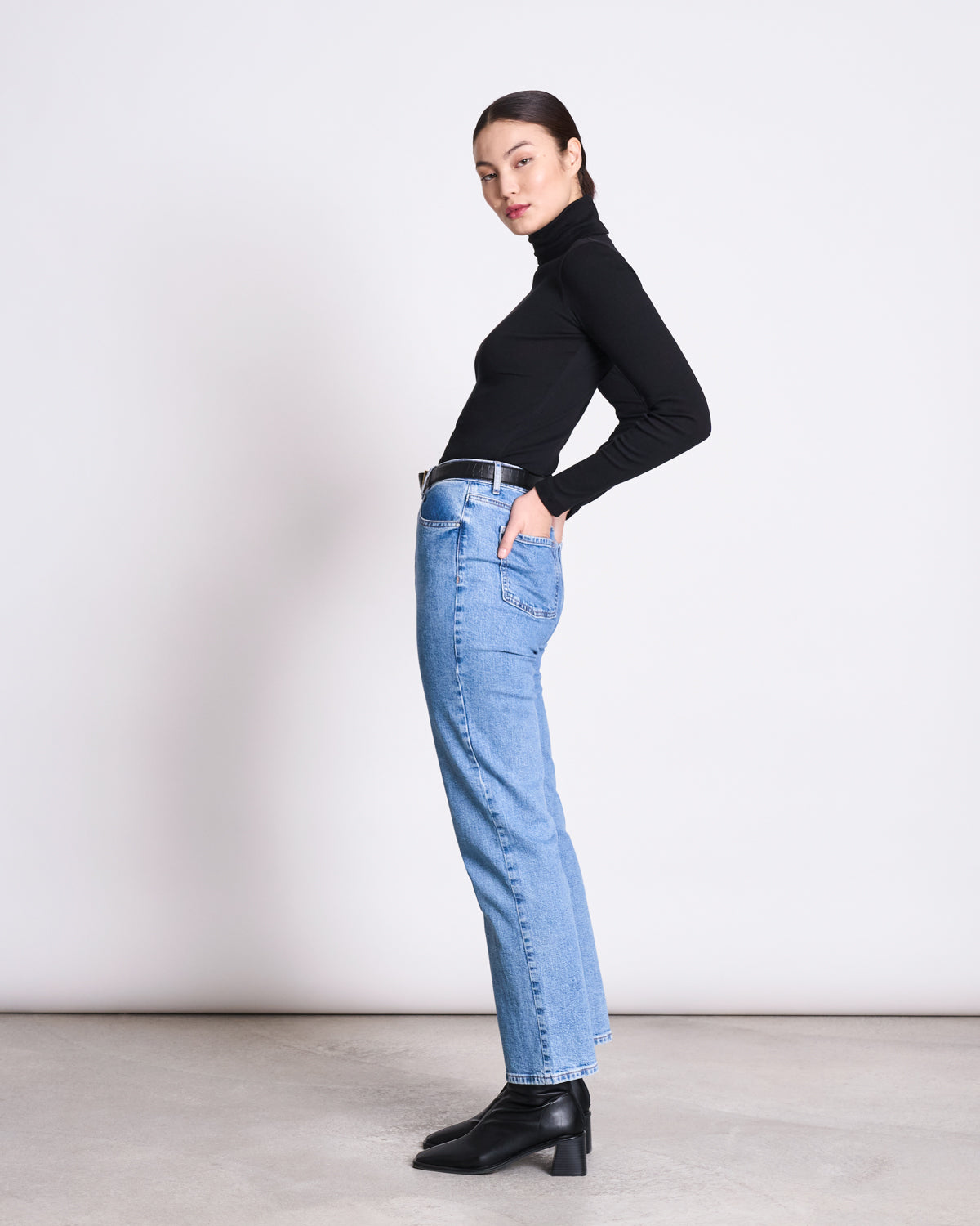 A person stands sideways against a plain light background, wearing the jan-n-june MINIRIB TURTLENECK CHUVA BLACK GOTS, light blue jeans, and black heeled boots, with hands in pockets and head slightly turned toward the camera.