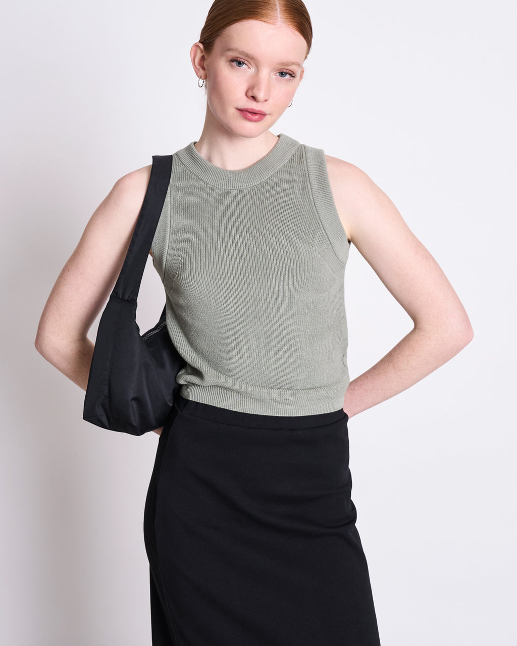 A red-haired woman with her hair tied back wears the jan-n-june KNIT TOP GARDA MUTED SEA SPRAY GOTS, a versatile gray-green sleeveless knit top. She pairs it with a black skirt and shoulder bag, posing against a plain light background.