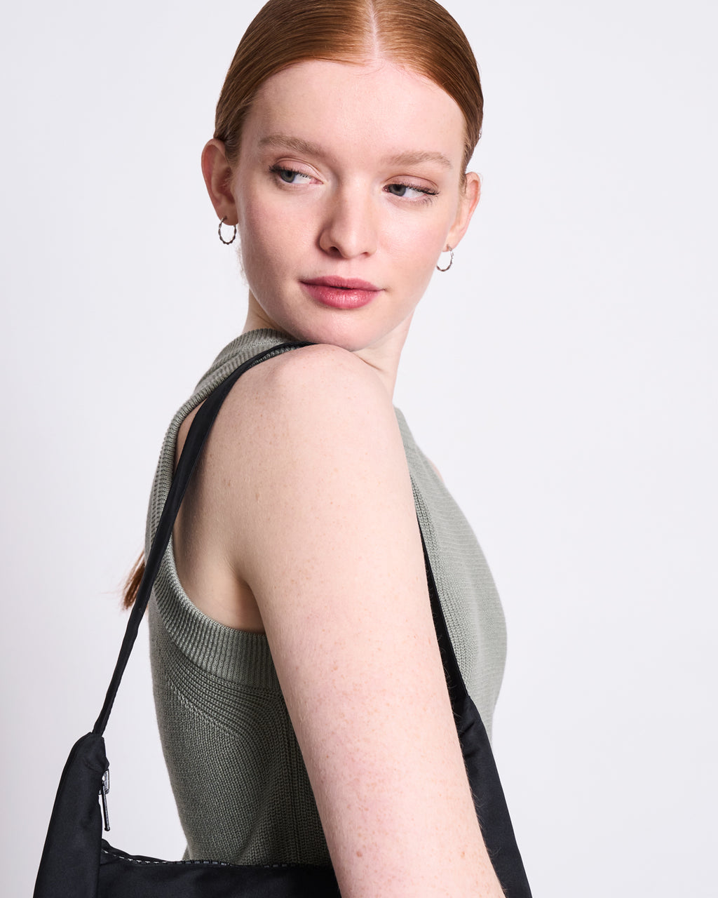 A woman with pale skin and straight red hair pulled back looks over her shoulder, wearing the jan-n-june KNIT TOP GARDA MUTED SEA SPRAY GOTS, small hoop earrings, and carrying a black bag against a plain light background.