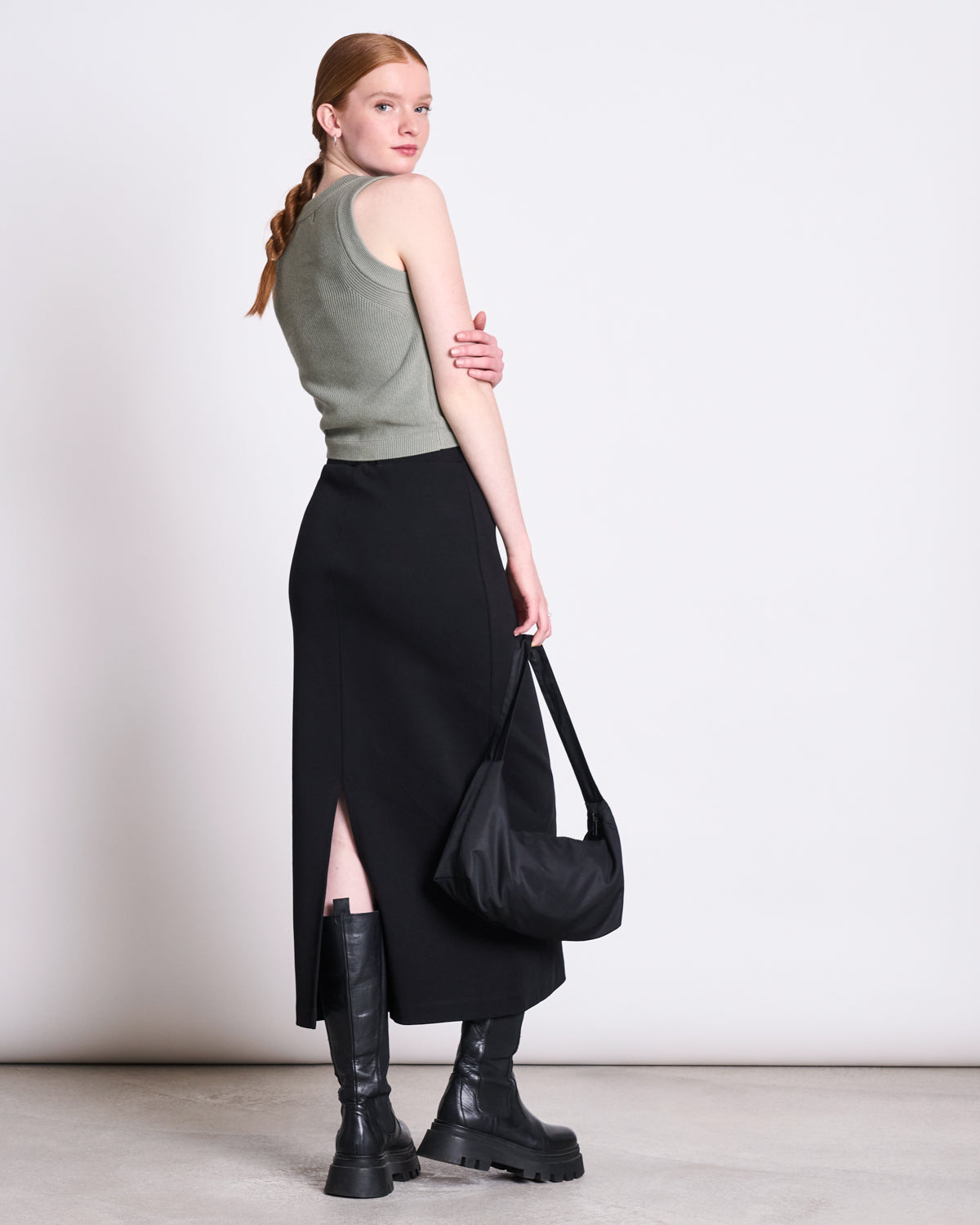A woman with long red braided hair wears the jan-n-june KNIT TOP GARDA MUTED SEA SPRAY GOTS and a black skirt, paired with chunky black boots and a black shoulder bag; she stands facing away, turning her head slightly over her shoulder.