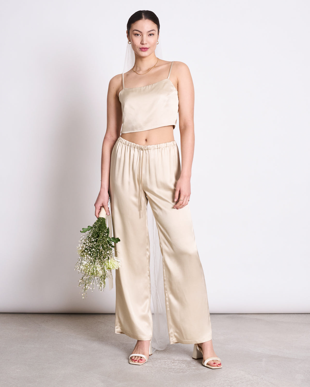 A woman in the jan-n-june BOW TOP LEEVKE IVORY and matching wide-leg pants, made from soft ENKA® Viscose, stands against a plain backdrop with a bouquet of flowers and open-toe heels.