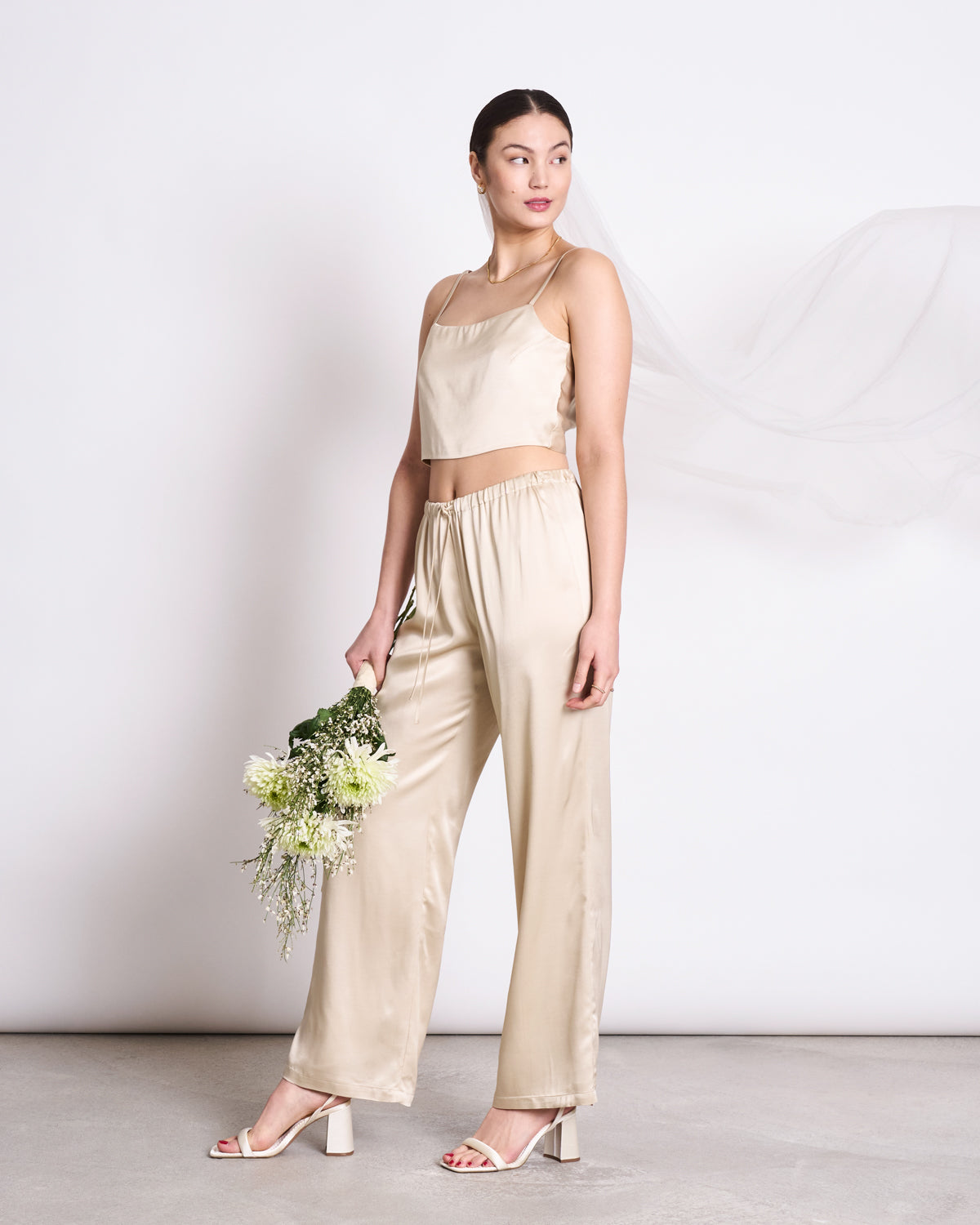 A woman in jan-n-june’s BOW TOP LEEVKE IVORY and matching wide-leg pants stands holding white flowers. She wears heeled sandals, her hair pulled back, with a flowing sheer ENKA® Viscose veil behind her.