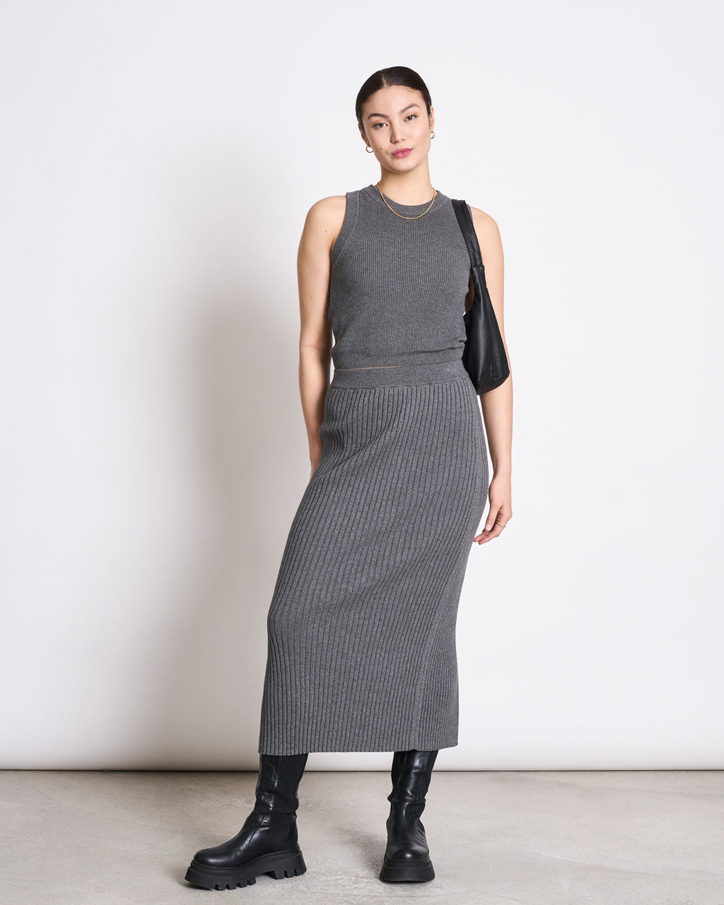 A woman wearing a sleeveless gray sweater top, the jan-n-june MIDI KNIT SKIRT EBBA DARK GREY GOTS with ribbed texture, black chunky boots, and a black shoulder bag stands against a plain white background.