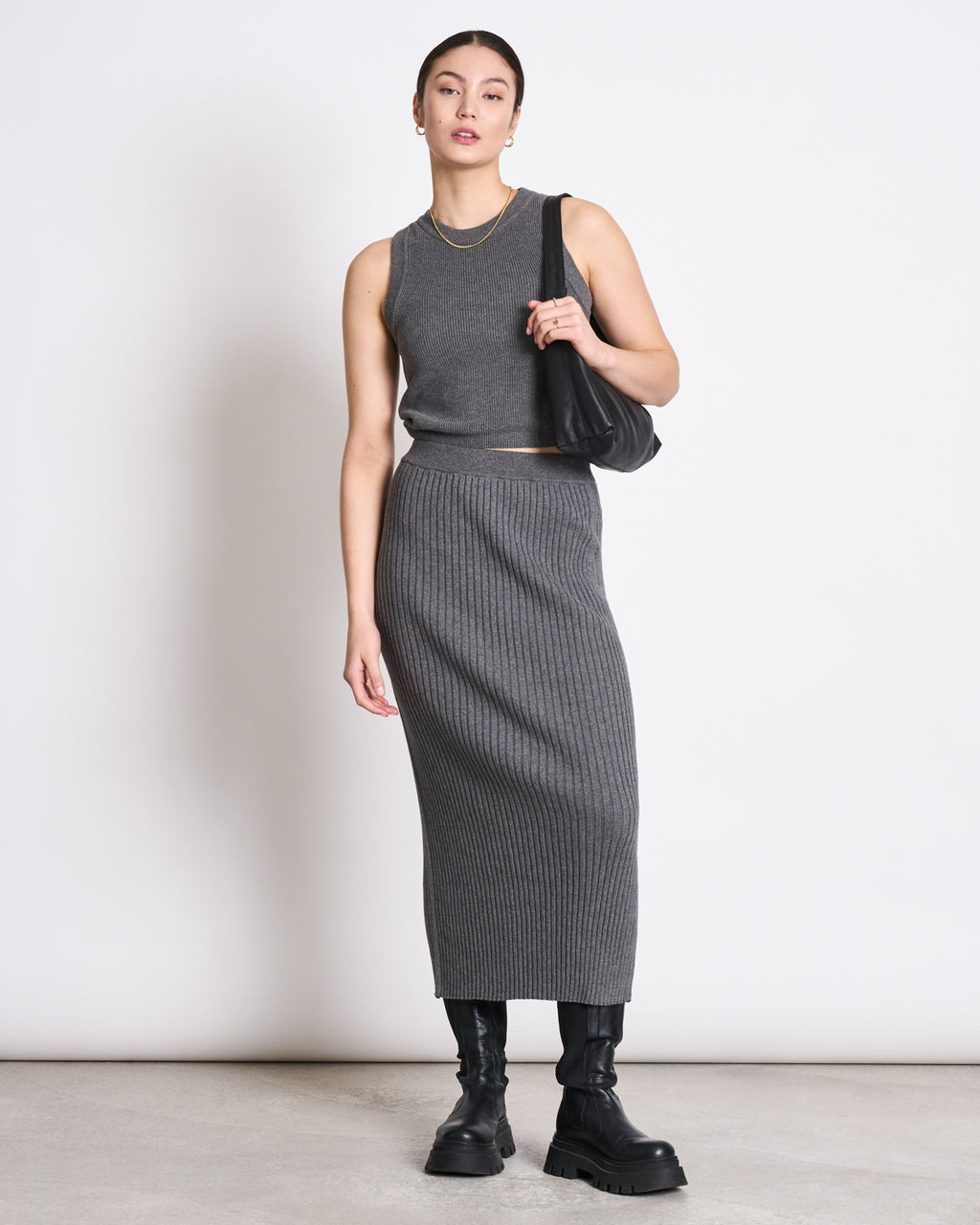 A woman stands against a plain background wearing a sleeveless gray top, the jan-n-june MIDI KNIT SKIRT EBBA DARK GREY GOTS with a ribbed texture, black chunky boots, and a black shoulder bag. Her hair is tied back and she accessorizes with a gold necklace.