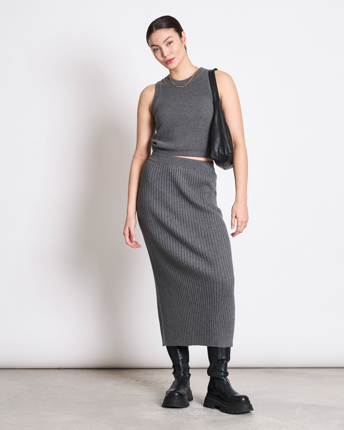 Against a plain white background, a woman wears the jan-n-june KNIT TOP GARDA DARK GREY GOTS with a matching ribbed gray midi skirt, chunky black boots, and carries a black shoulder bag. Her hair is tied back and she accessorizes with hoop earrings.