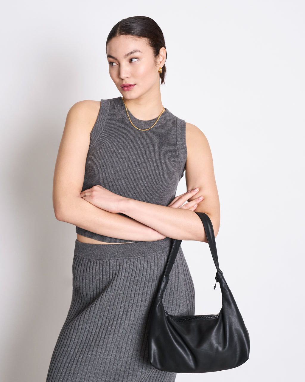A woman stands with arms crossed, holding a black handbag and wearing the jan-n-june KNIT TOP GARDA DARK GREY GOTS—an organic cotton sleeveless top—looking to the side against a plain white background, highlighting its versatile style.