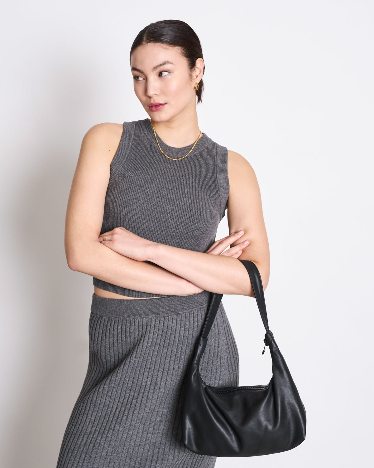 A woman stands with arms crossed, holding a black handbag and wearing the jan-n-june KNIT TOP GARDA DARK GREY GOTS—an organic cotton sleeveless top—looking to the side against a plain white background, highlighting its versatile style.