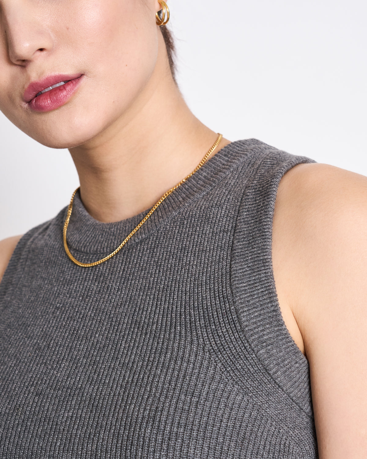 A person models the jan-n-june KNIT TOP GARDA DARK GREY GOTS—a versatile, sleeveless ribbed knit—styled with a slim gold necklace and gold hoop earring, shown from the shoulders up against a plain white background.