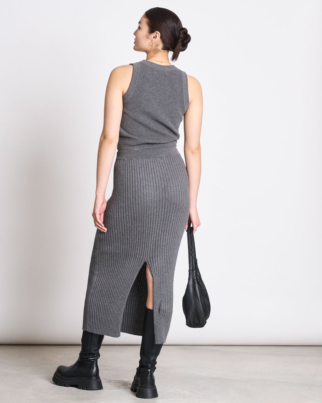 A woman with a low bun wears the jan-n-june KNIT TOP GARDA DARK GREY GOTS, an organic cotton sleeveless knit, paired with a ribbed gray midi skirt with back slit, black bag and boots for a versatile look.