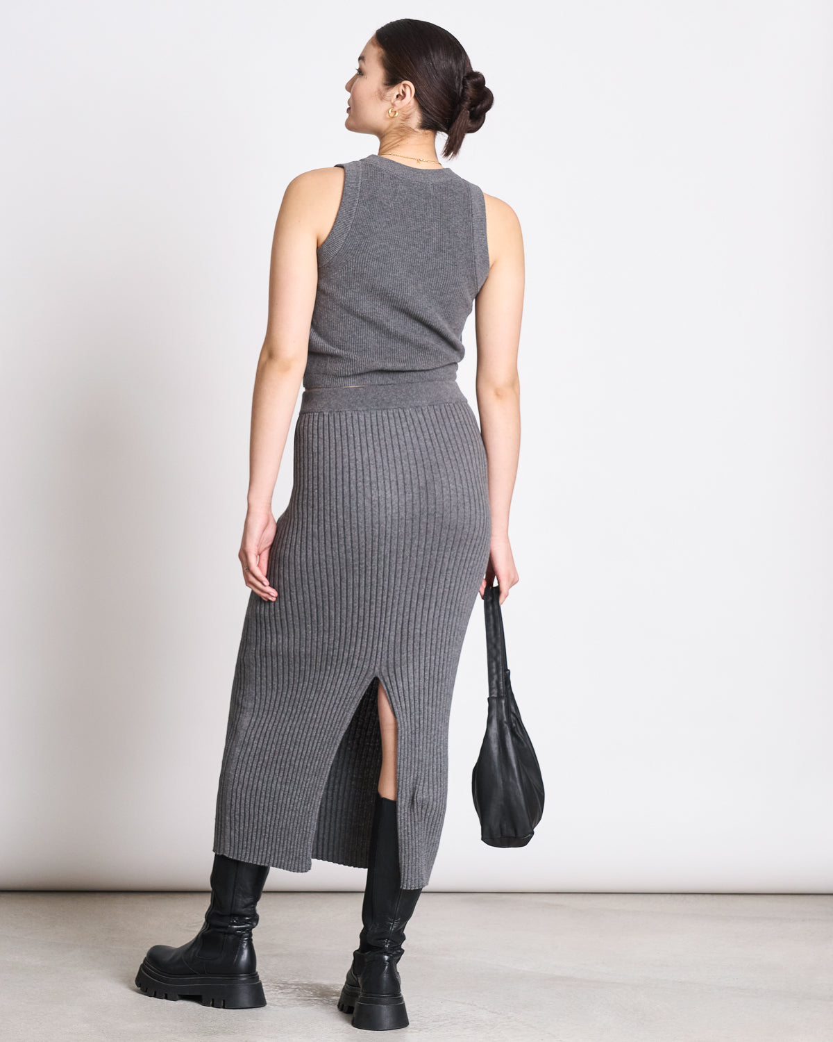 A woman with dark hair in a bun faces away, wearing jan-n-june’s sleeveless gray knit top and the MIDI KNIT SKIRT EBBA DARK GREY GOTS, both with ribbed texture and a back slit, paired with black boots and a handbag.