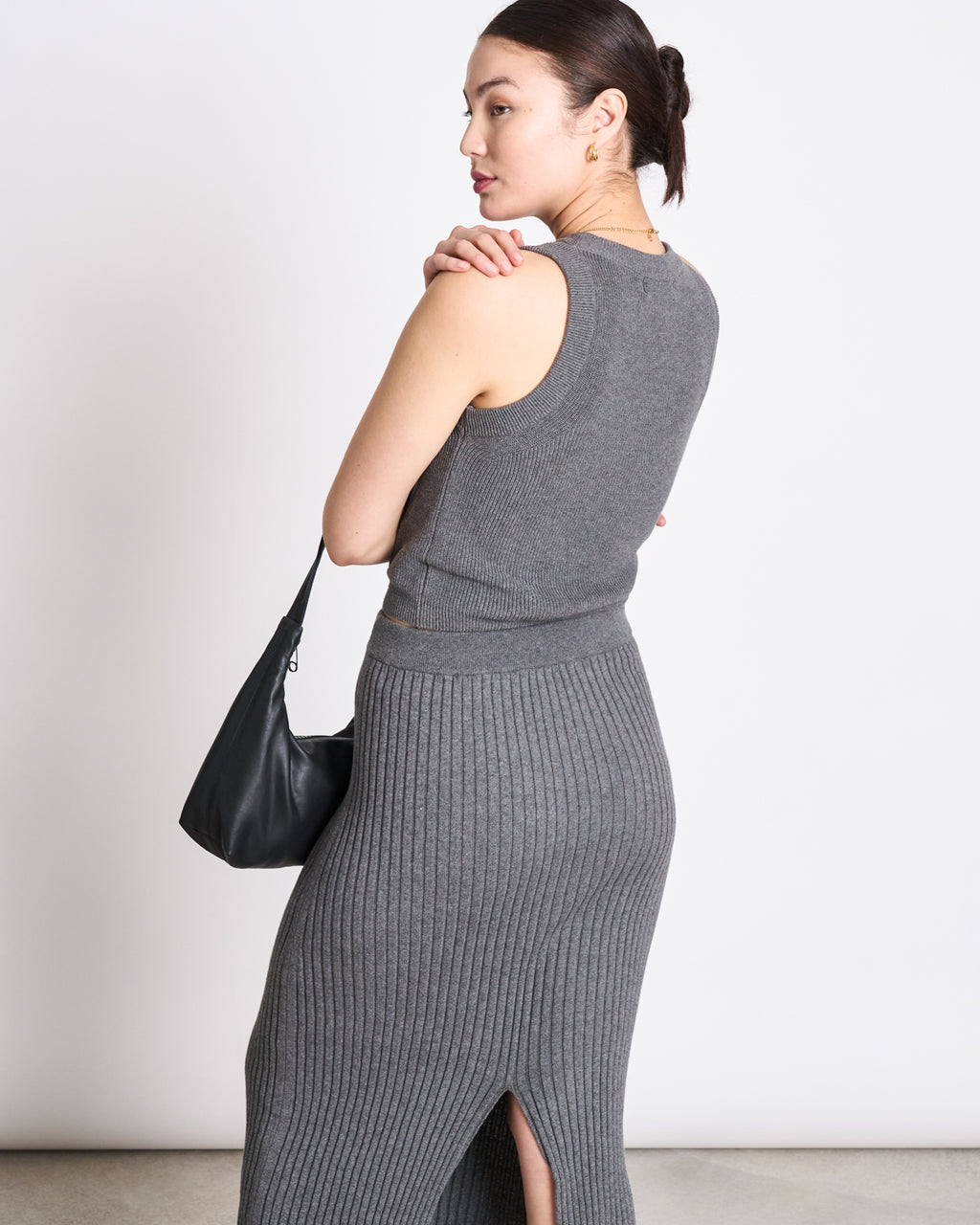 A woman faces away, wearing the jan-n-june KNIT TOP GARDA DARK GREY GOTS and a matching high-waisted skirt with a back slit. She carries a black shoulder bag; her dark hair is tied back against a plain white background.