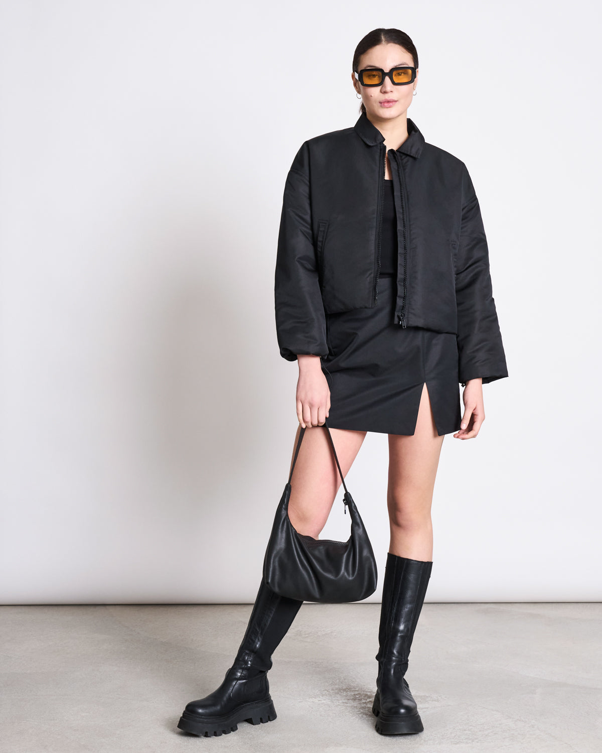 A woman stands against a plain background wearing the jan-n-june MINI REC. SKIRT TAZE BLACK, a black cropped jacket, knee-high black boots, sunglasses, and holding a black handbag.