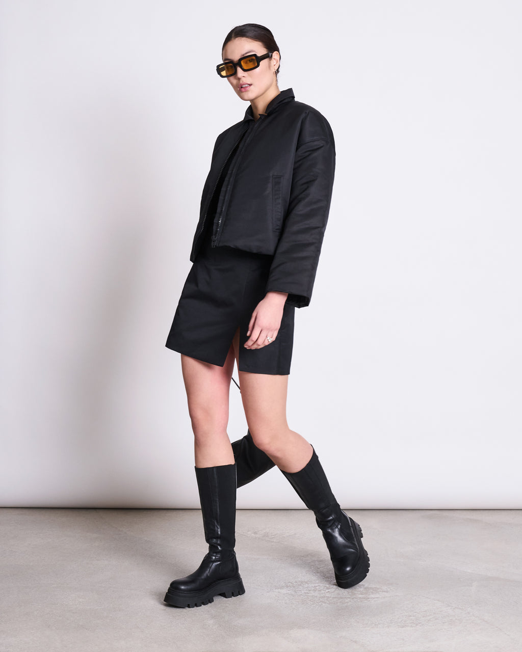 A person stands against a light wall wearing the jan-n-june REC. BLOUSON DUSK BLACK made from recycled material, paired with a black mini skirt, knee-high black boots, large brown sunglasses, hair pulled back, and one hand by their side.
