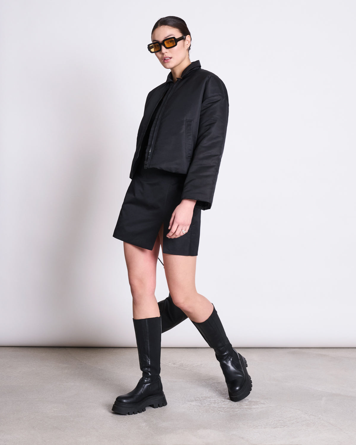 A person stands against a light wall wearing the jan-n-june REC. BLOUSON DUSK BLACK made from recycled material, paired with a black mini skirt, knee-high black boots, large brown sunglasses, hair pulled back, and one hand by their side.