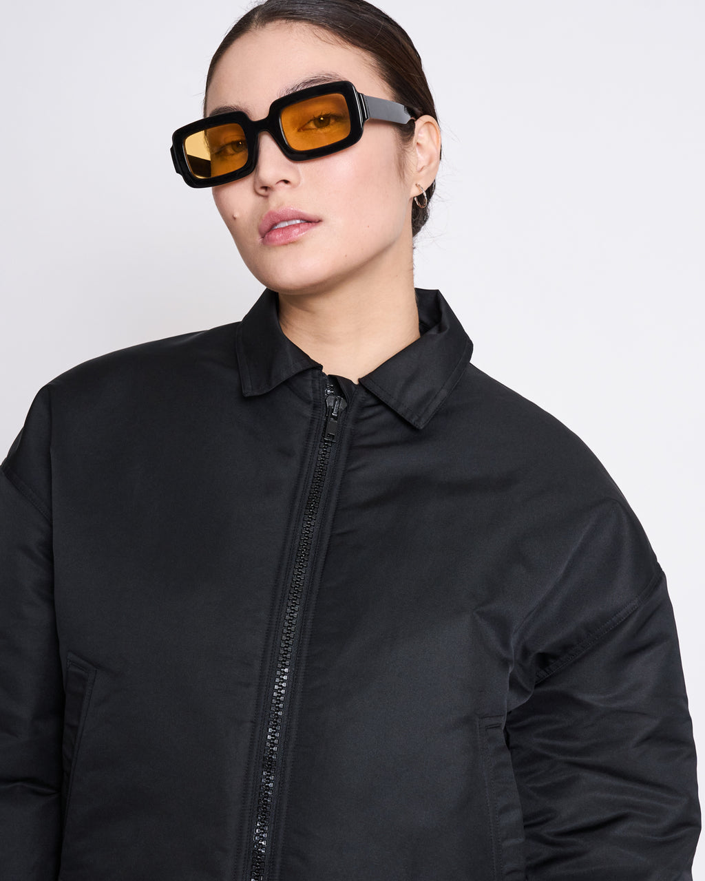 A woman wears jan-n-junes REC. BLOUSON DUSK BLACK, a sustainable jacket made from recycled material, paired with black square sunglasses with orange lenses, standing against a plain light background.