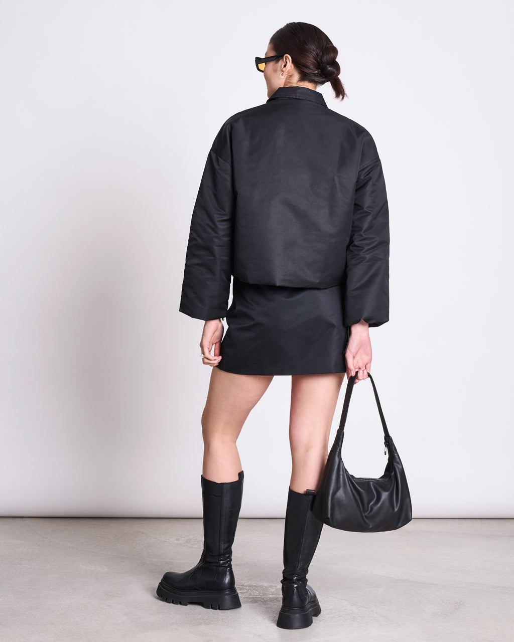 A woman with dark hair in a low bun stands facing away against a plain white background, wearing the jan-n-june REC. BLOUSON DUSK BLACK jacket, black mini skirt, knee-high boots, sunglasses, and holding a black handbag.