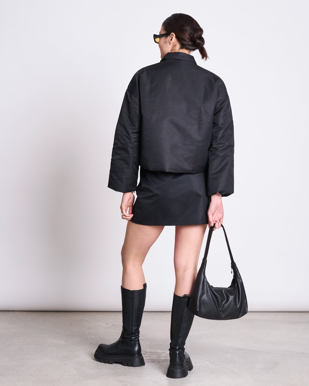 A woman with her hair in a low bun wears jan-n-junes MINI REC. SKIRT TAZE BLACK, paired with a matching jacket, knee-high boots, sunglasses, and carries a black handbag—embodying chic sustainable fashion.