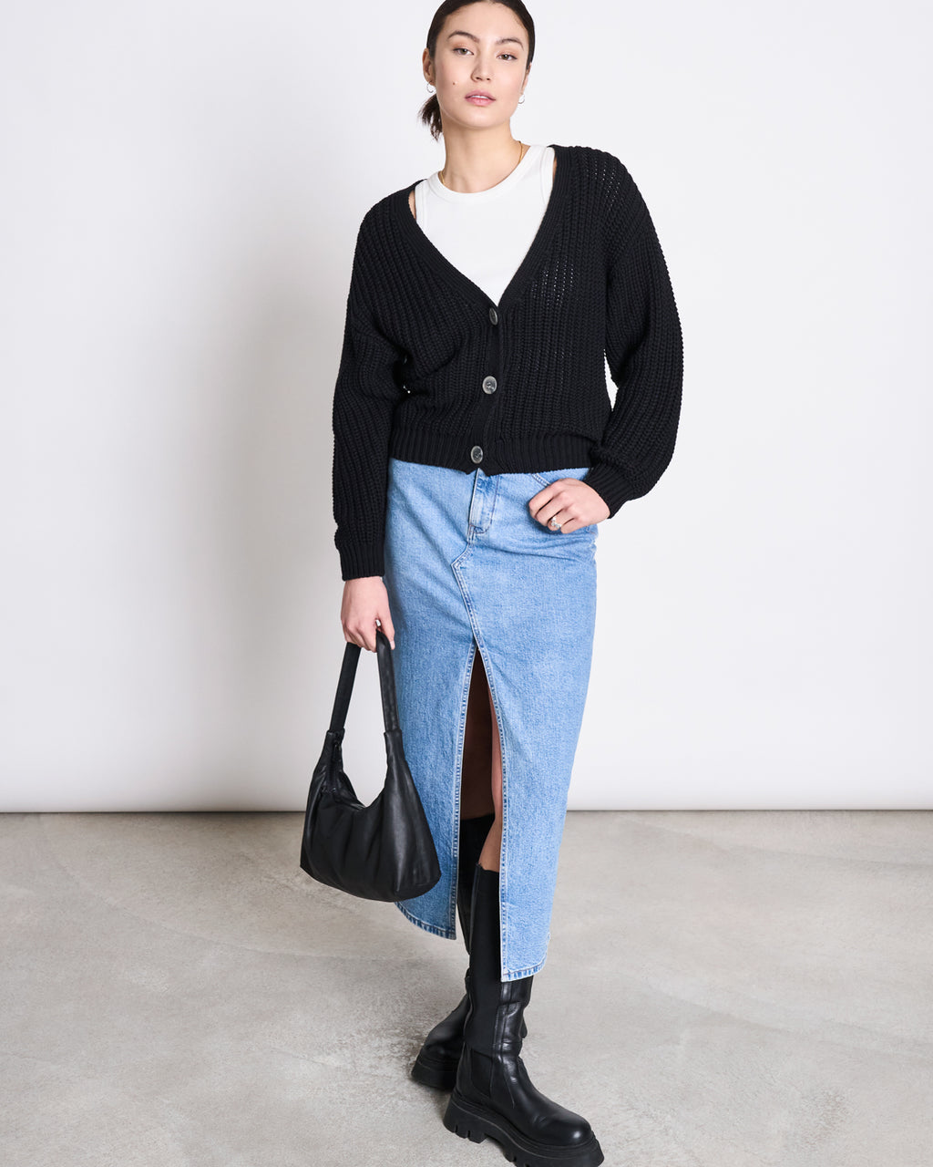 A woman stands against a plain background wearing the jan-n-june CHUNKY CARDIGAN LENA BLACK GOTS—her bestseller look in 100% organic cotton—styled with a white shirt, light blue denim skirt with front slit, black chunky boots, and black handbag.