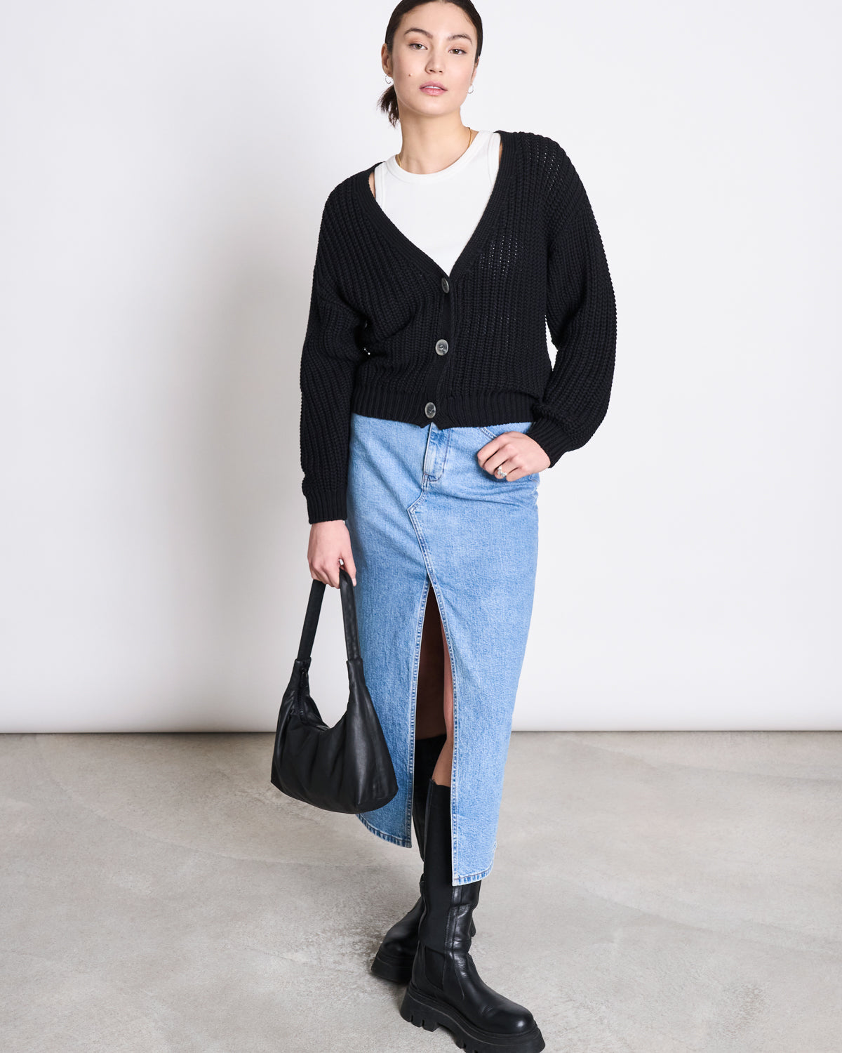 A woman stands against a plain background wearing the jan-n-june CHUNKY CARDIGAN LENA BLACK GOTS—her bestseller look in 100% organic cotton—styled with a white shirt, light blue denim skirt with front slit, black chunky boots, and black handbag.