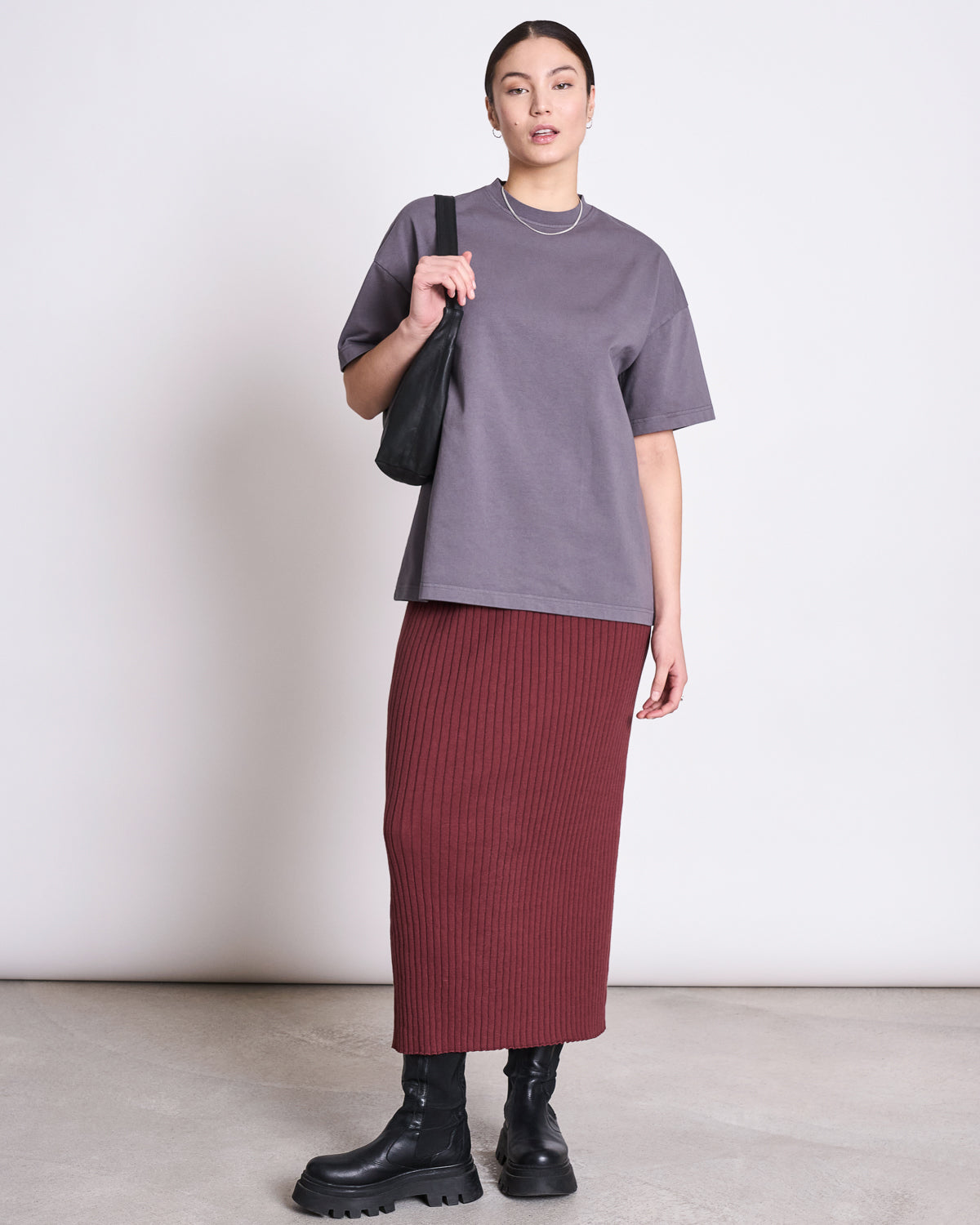 A person stands against a plain background wearing the jan-n-june MIDI KNIT SKIRT EBBA INTENSE RUST GOTS, a loose gray T-shirt, chunky black boots, and a black shoulder bag. Their hair is pulled back as they look at the camera.