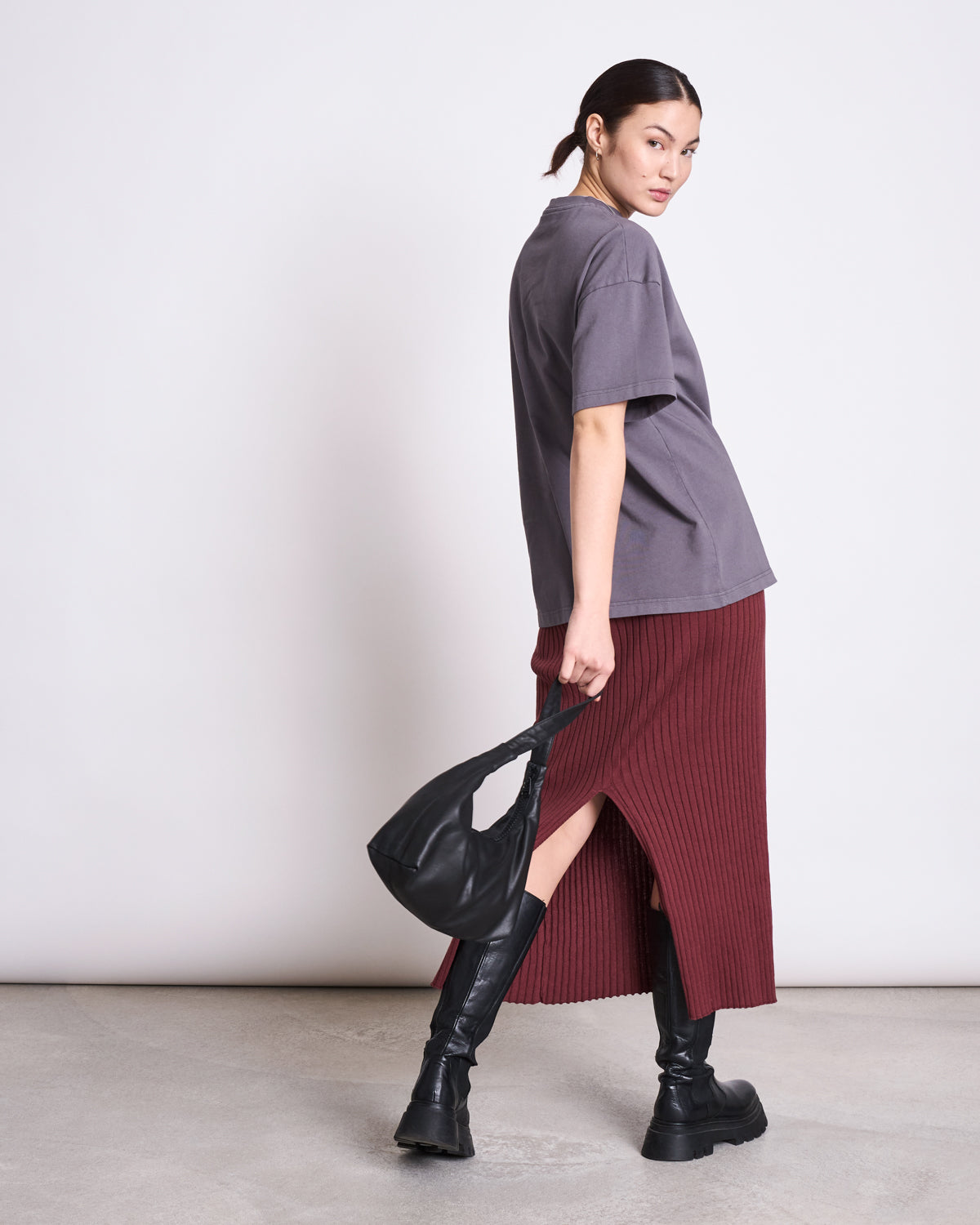 A woman stands sideways, looking over her shoulder. She wears a loose gray T-shirt, black knee-high boots, holds a black bag, and the jan-n-june MIDI KNIT SKIRT EBBA INTENSE RUST GOTS. The background is plain and light-colored.