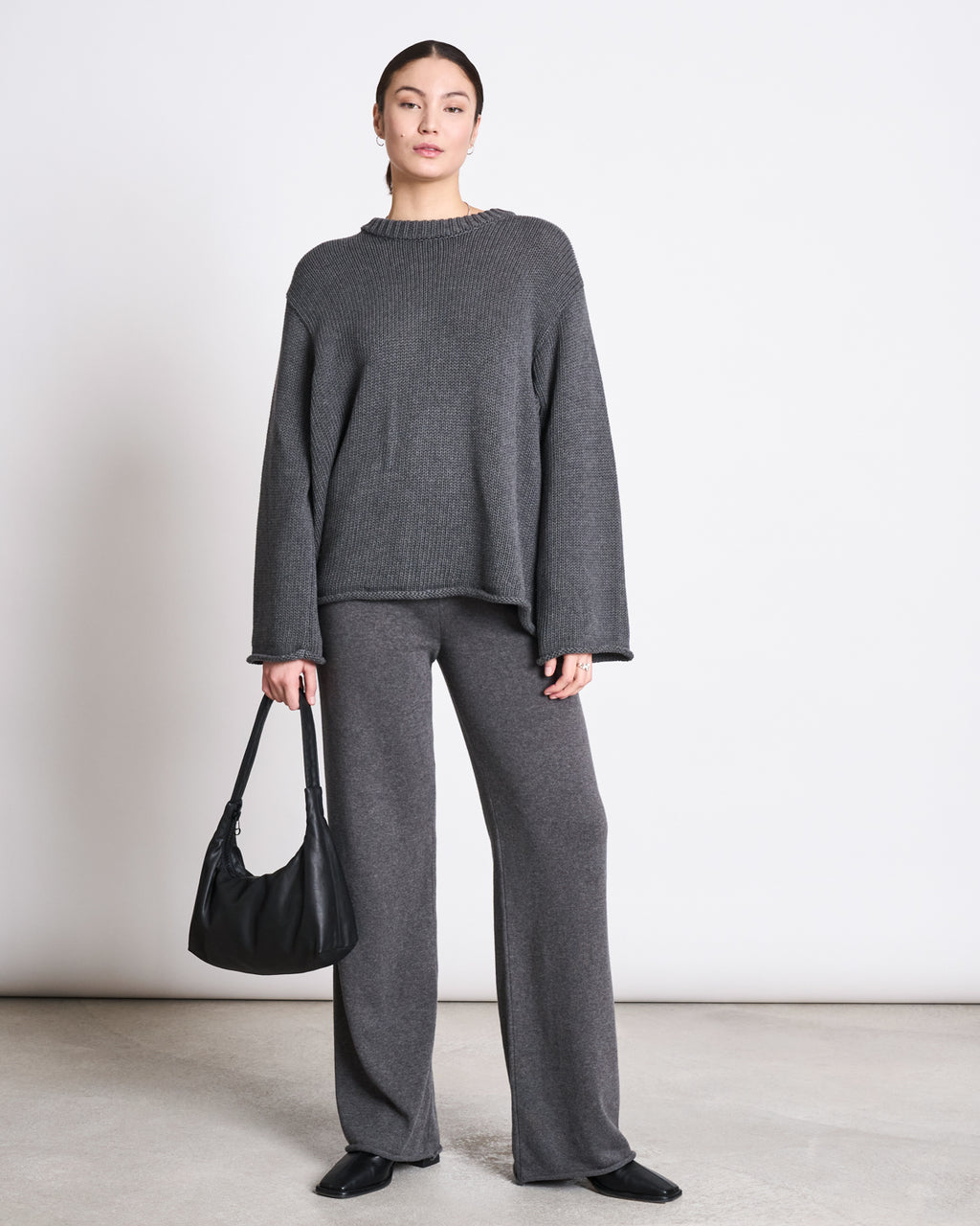 A woman stands against a plain white wall wearing the jan-n-june JUMPER SONA DARK GREY GOTS, styled with matching wide-leg pants. She holds a black handbag, wears black shoes, and has her hair pulled back.