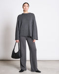 A woman stands against a plain white wall wearing the jan-n-june JUMPER SONA DARK GREY GOTS, styled with matching wide-leg pants. She holds a black handbag, wears black shoes, and has her hair pulled back.