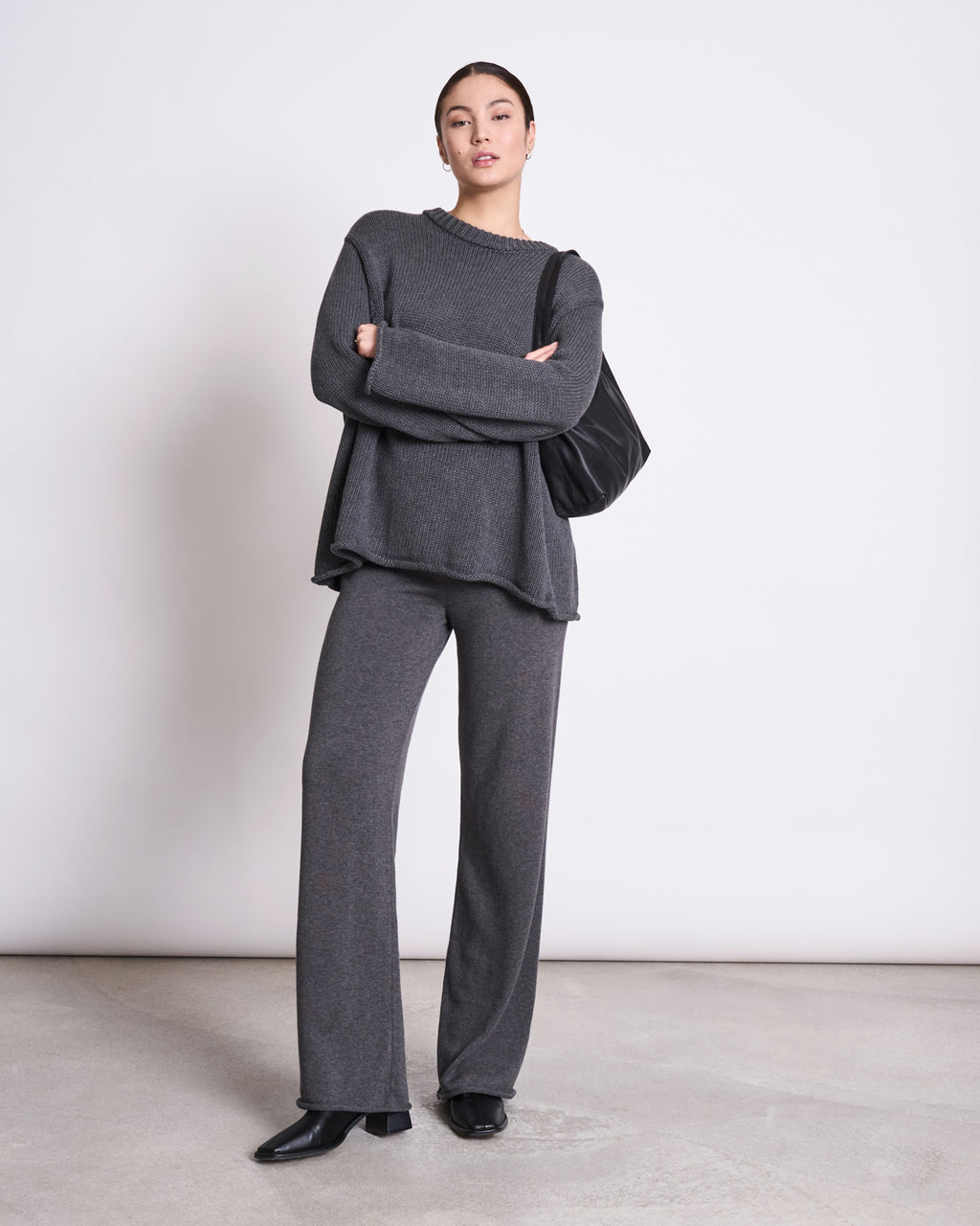A woman poses confidently against a plain wall in the jan-n-june JUMPER SONA DARK GREY GOTS—an oversized, GOTS certified organic cotton sweater—paired with matching wide-leg pants, black boots, and a black shoulder bag.