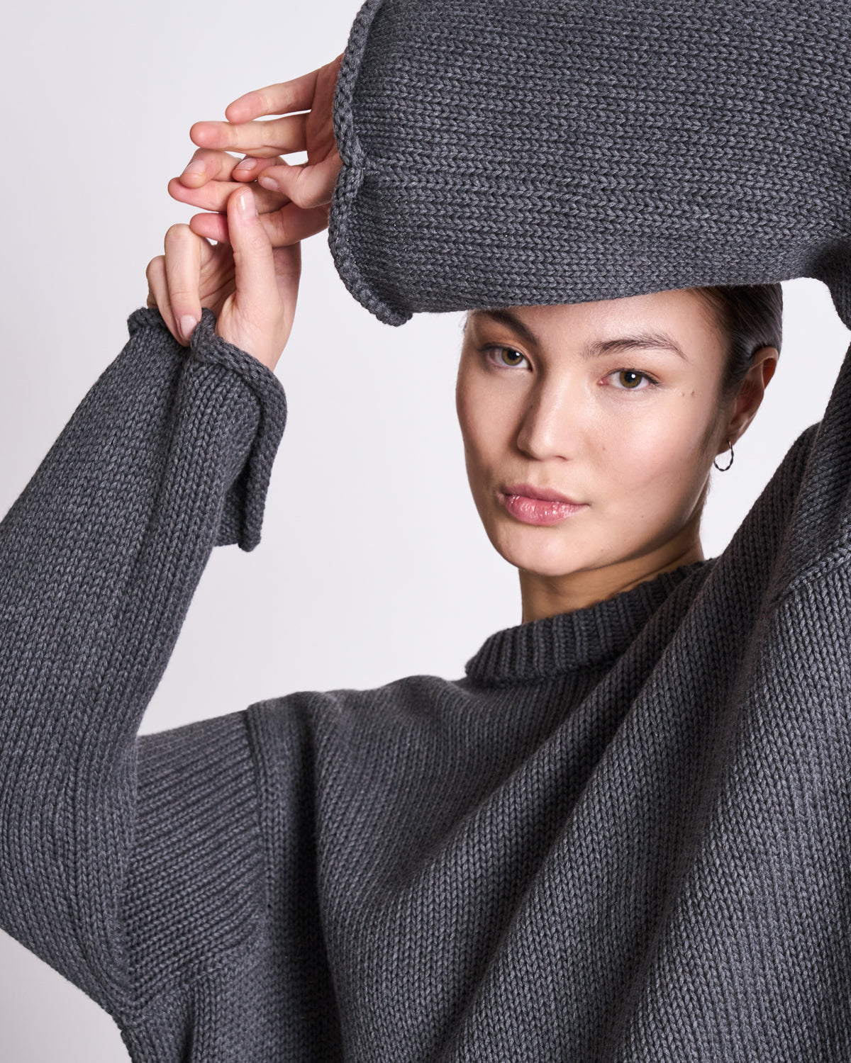 A person wearing the jan-n-june JUMPER SONA DARK GREY GOTS sweater raises one arm, partly covering their forehead, and gazes neutrally at the camera against a light background.