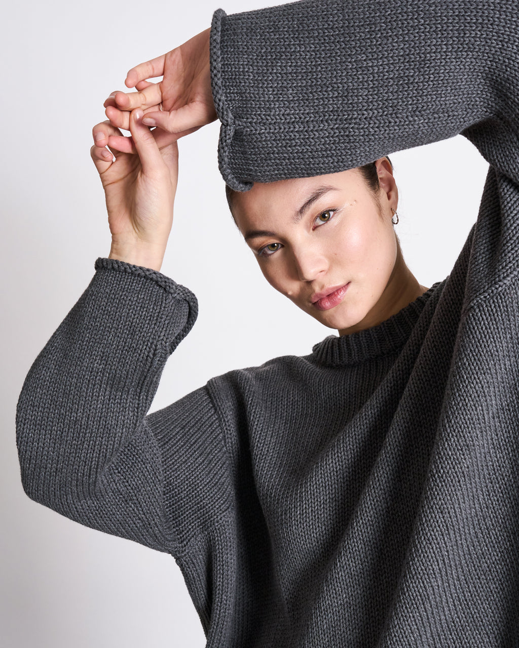 A person wears the jan-n-june JUMPER SONA DARK GREY GOTS, an oversized organic cotton sweater, posing with arms raised and a neutral expression against a plain backdrop.