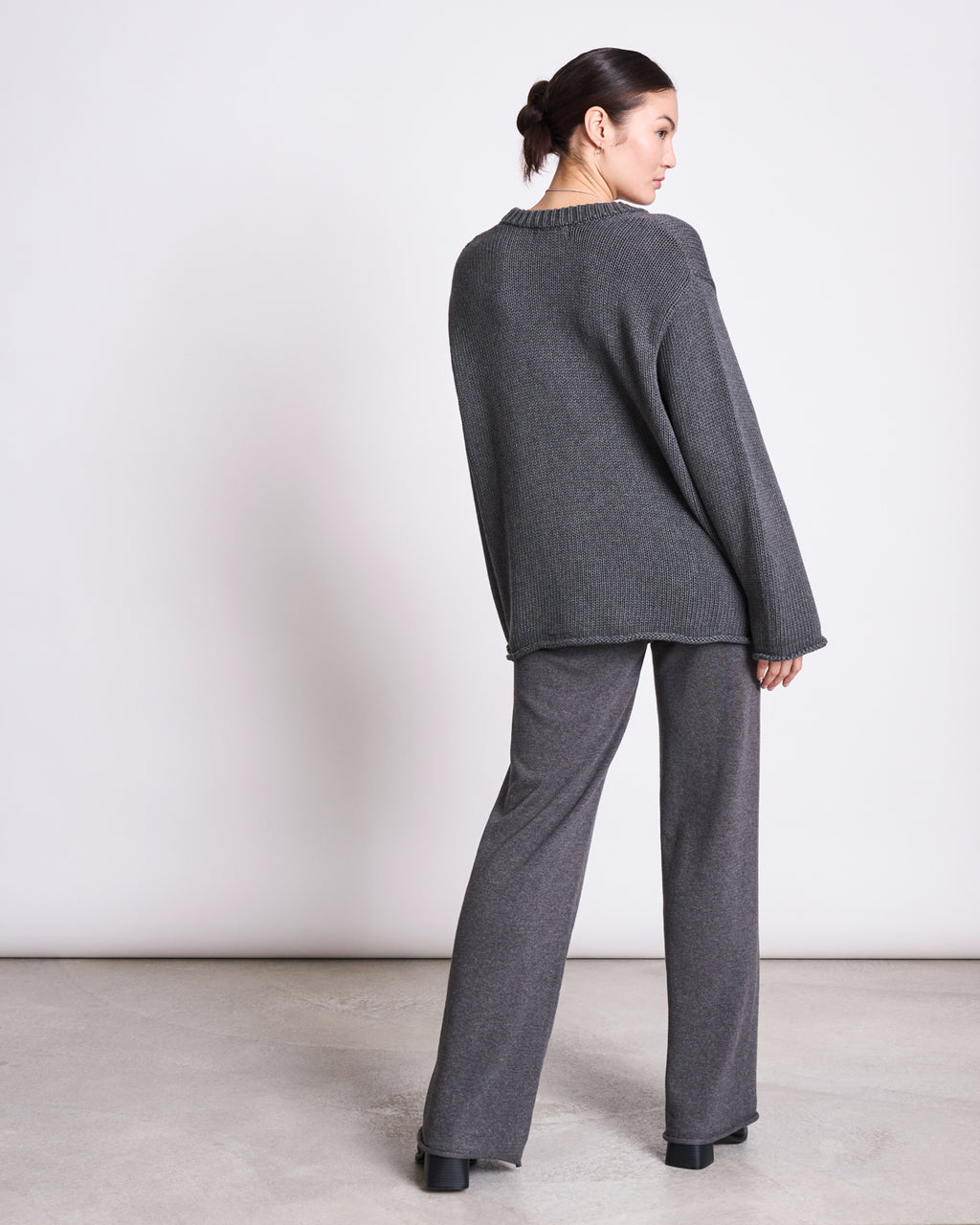 A woman faces away, wearing the jan-n-june JUMPER SONA DARK GREY GOTS—an oversized dark gray sweater with wide-leg pants and black platform shoes. Her hair is in a low bun against a plain, light-colored background.
