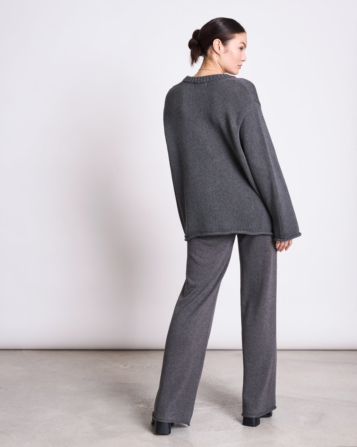 A woman faces away, wearing the jan-n-june JUMPER SONA DARK GREY GOTS—an oversized dark gray sweater with wide-leg pants and black platform shoes. Her hair is in a low bun against a plain, light-colored background.