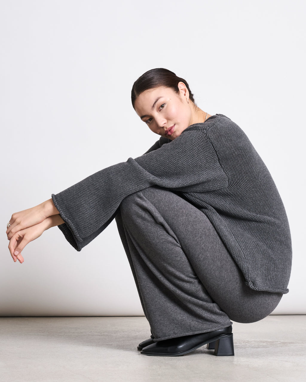 A woman with dark hair pulled back squats on the floor, wearing the jan-n-june JUMPER SONA DARK GREY GOTS—a loose, organic cotton sweater—paired with soft gray pants and black heeled shoes. She looks downward calmly against a plain light background.