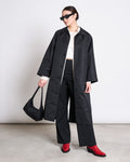 A woman in the jan-n-june REC. CAR COAT HAZE BLACK—crafted with SEAQUAL® YARN—pairs it with black pants, a white top, red boots, sunglasses, and a black bag, standing against a plain background and showcasing sustainable style.
