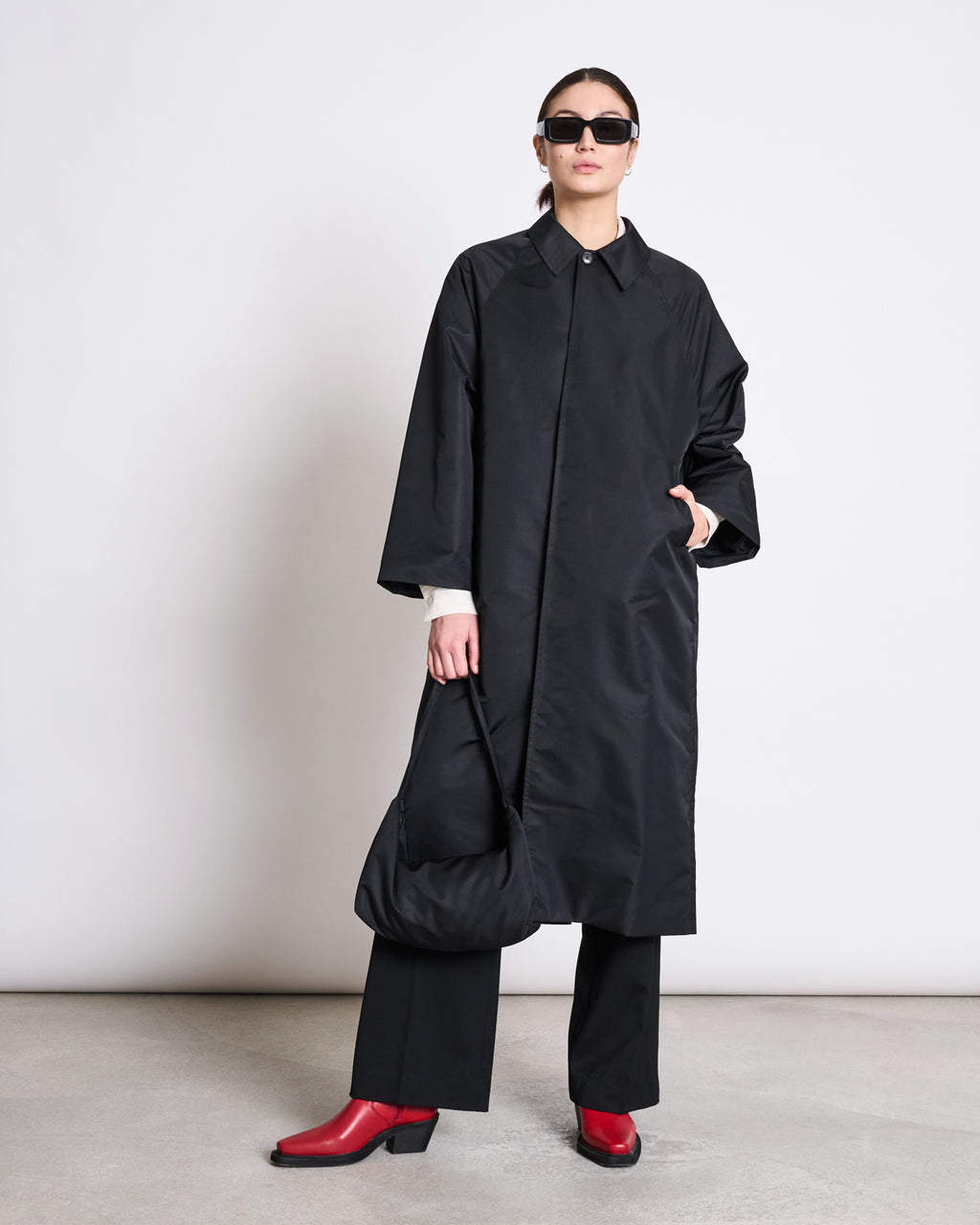 A person in the jan-n-june REC. CAR COAT HAZE BLACK made from SEAQUAL® YARN, paired with black pants, red heeled boots, sunglasses, and a black bag against a plain background—sustainable fashion at its best.