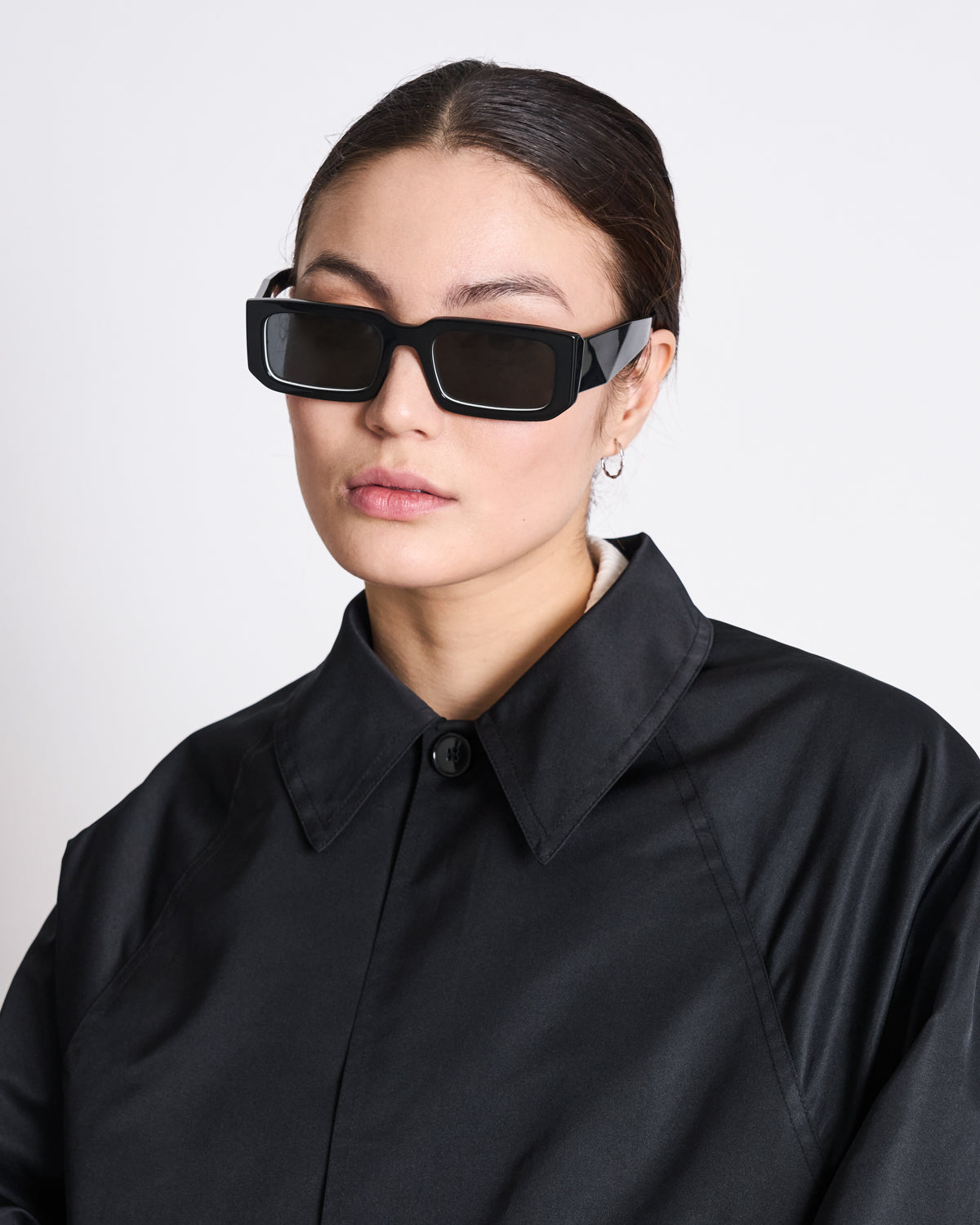 A woman with dark hair tied back wears black rectangular sunglasses, small hoop earrings, and the jan-n-june REC. CAR COAT HAZE BLACK made from SEAQUAL® YARN, standing against a plain white background.