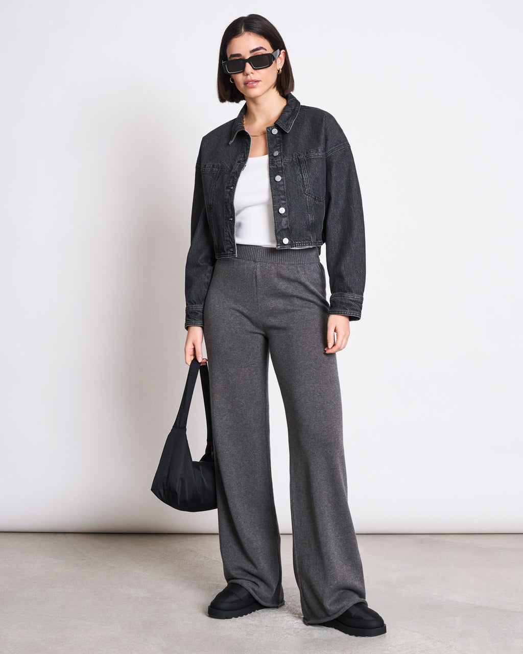 A woman in sunglasses wears the jan-n-june DENIM JACKET ARIA FADED BLACK over a white top, pairs it with loose gray pants and black shoes, and stands against a plain background holding a black bag in her left hand.