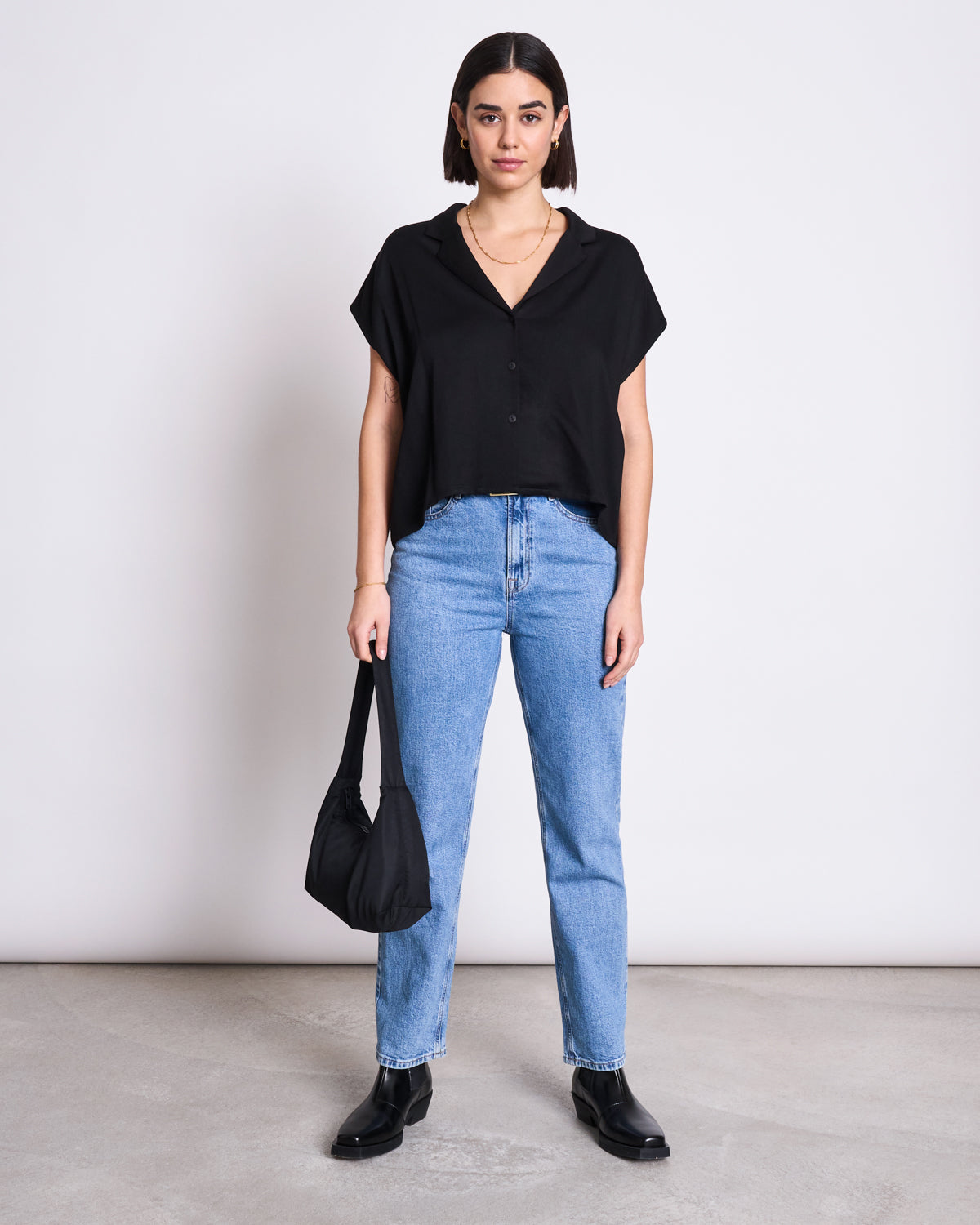 A woman faces forward against a plain background, wearing the jan-n-june BLOUSE TIERRA BLACK, light blue high-waisted jeans, black ankle boots, and holding a black handbag.
