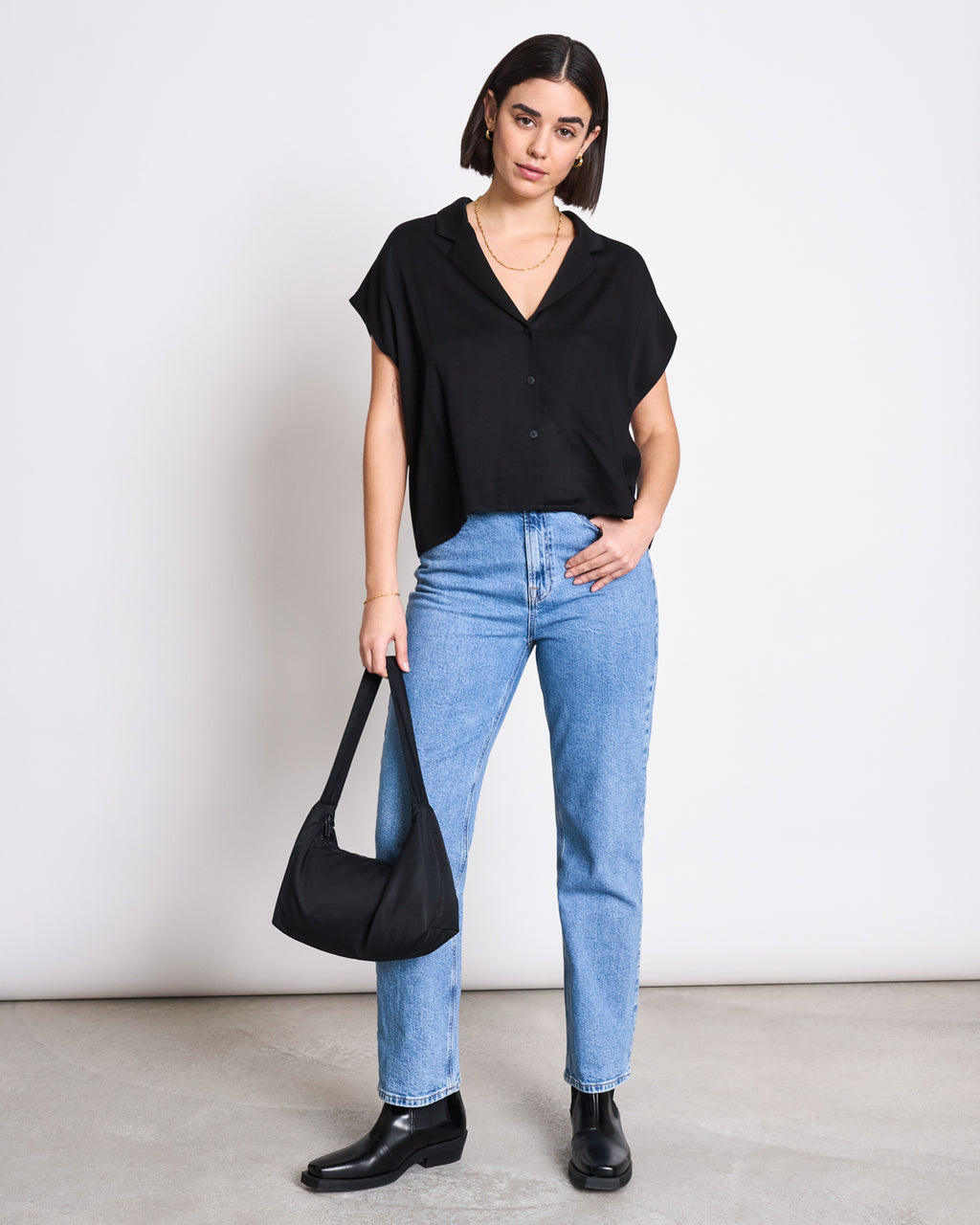 A woman stands against a plain background wearing the jan-n-june BLOUSE TIERRA BLACK, light blue high-waisted jeans, black ankle boots, and holding a black handbag.