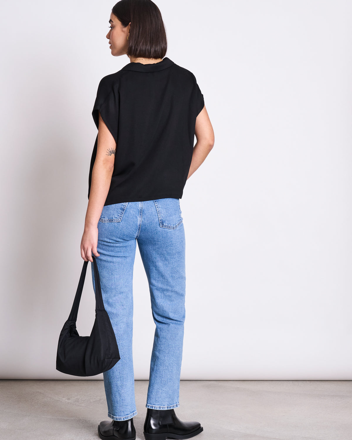 A person with straight dark hair stands facing away, wearing the jan-n-june BLOUSE TIERRA BLACK, light blue jeans, black ankle boots, and holding a black handbag against a plain light-colored background.