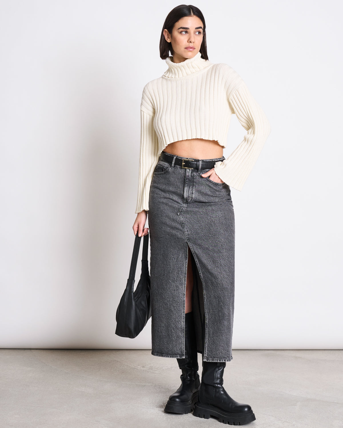 A woman stands against a white background wearing a cream ribbed turtleneck crop sweater, a jan-n-june DENIM SKIRT LUCE LIGHT GREY with a front slit, black boots, and holding a black handbag.