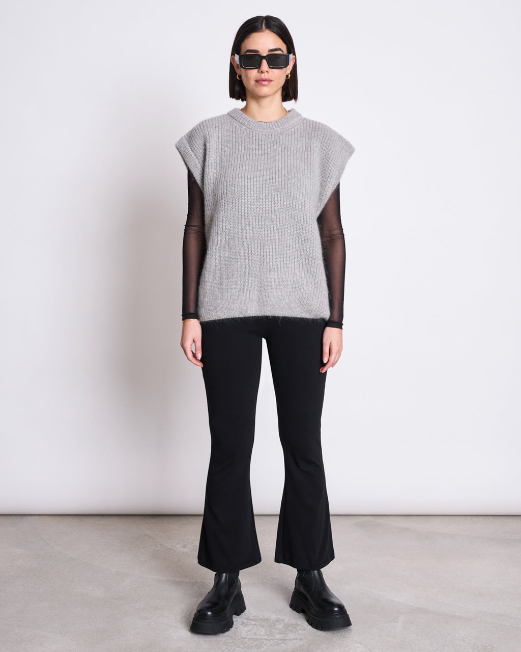 A person stands against a plain white wall wearing black sunglasses, the jan-n-june WOOLY SLIPOVER THANDA LIGHT GREY sweater over a black long-sleeve top, paired with black flared pants and chunky boots.